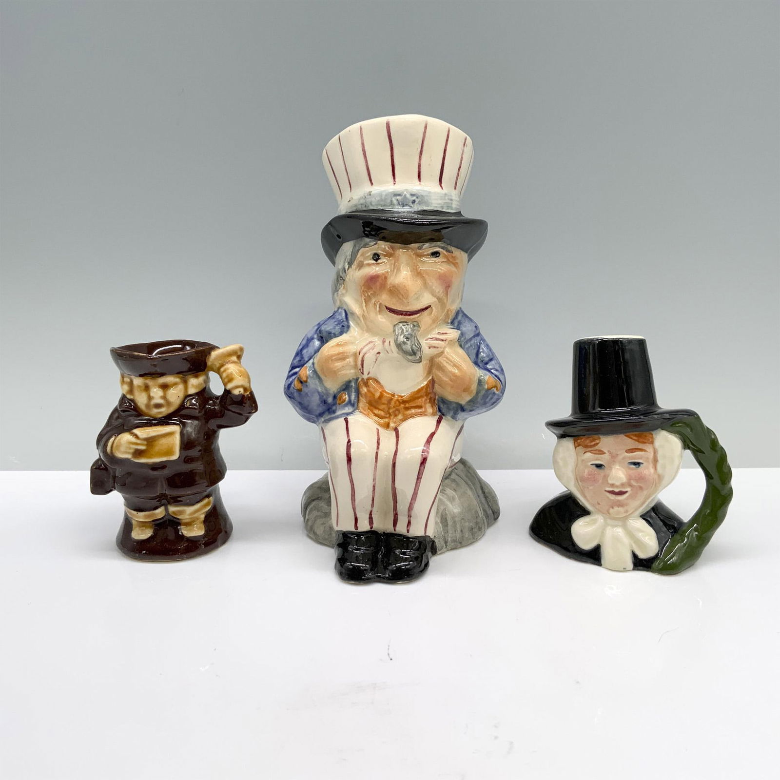 3pc Shorter and Son & American Themed Character & Toby Jugs: A set of three Toby jugs, one depicting Uncle Sam in a festive red, white, and blue striped outfit, another featuring a town crier dressed in brown with a bell and envelope, and a character jug showca