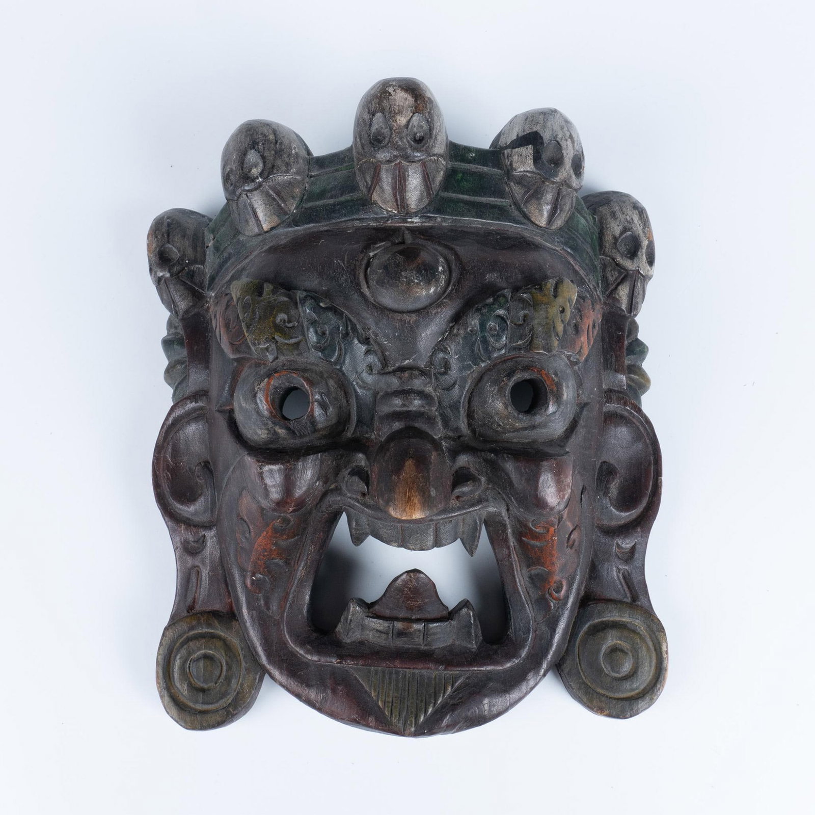 Tibetan Mahakala Carved Wooden Mask: Heavily carved symbolic mask with heads along top and wide-open mouth with fierce fanged teeth. Issued: 20th century Dimensions: 12"L x 10"W x 5"H Country of Origin: Tibet