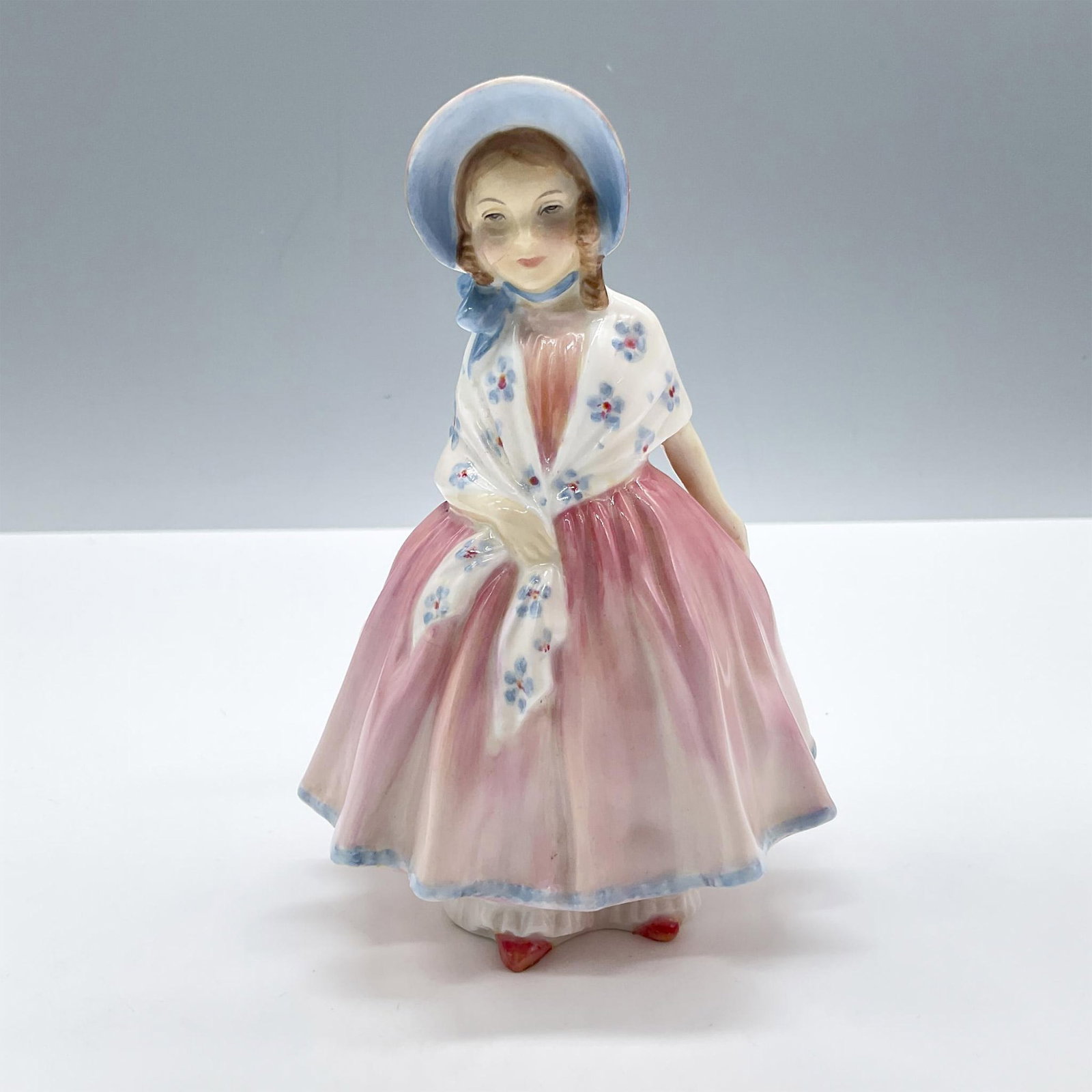 Lily HN1798 - Royal Doulton Figurine: Glossy porcelain. Features a lovely little girl wearing a soft pink dress and a white shawl with blue flowers. Royal Doulton backstamp. Artist: Leslie Harradine Issued: 1936 - 1971 Dimensions: 2.75''L