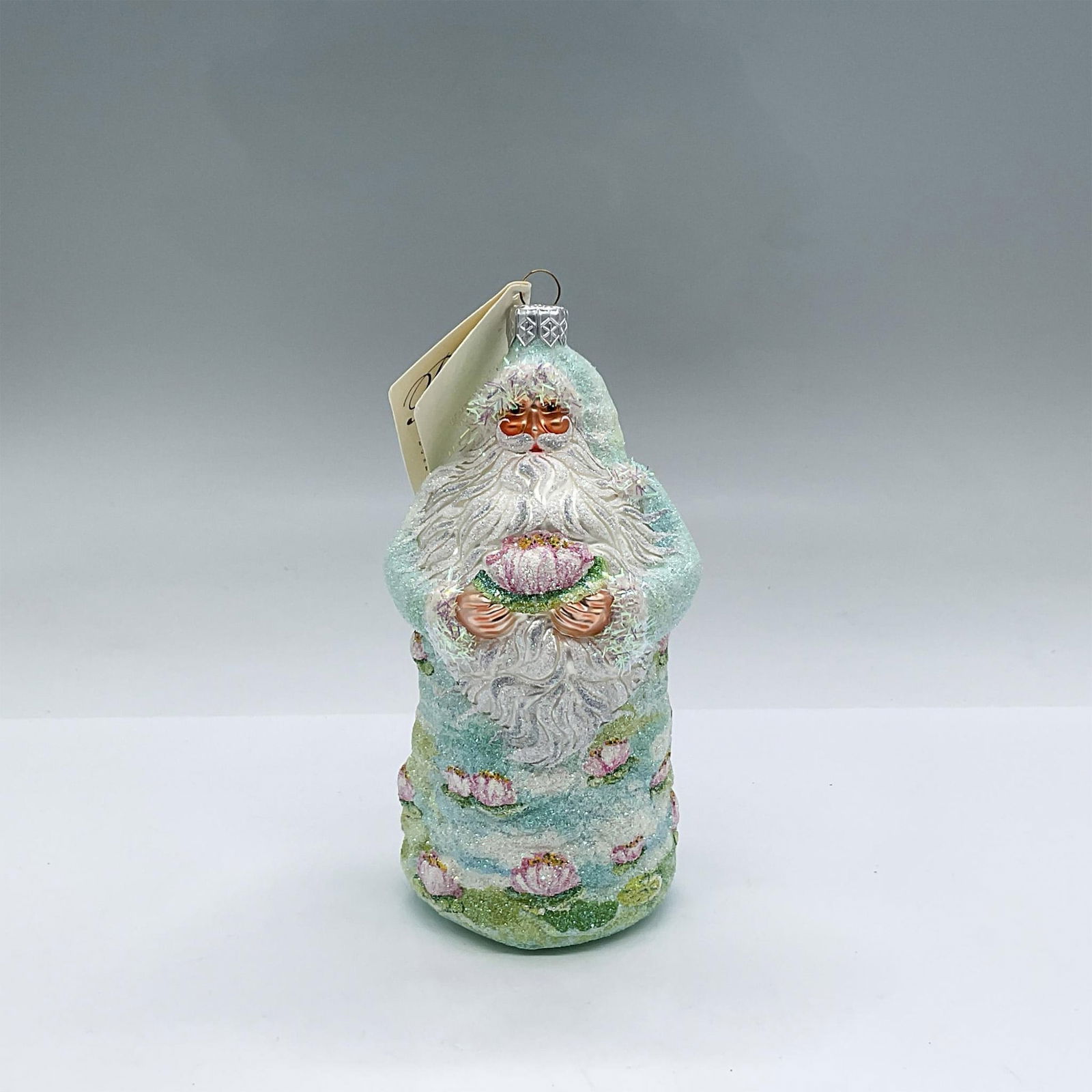 Patricia Breen Christmas Ornament, Lotus Santa: Exquisitely hand-painted in a European style, this glass ornament features Santa clad in a sky blue and green outfit adorned with lotuses. He is holding a pink lotus with both hands. Dimensions: 2.5"L