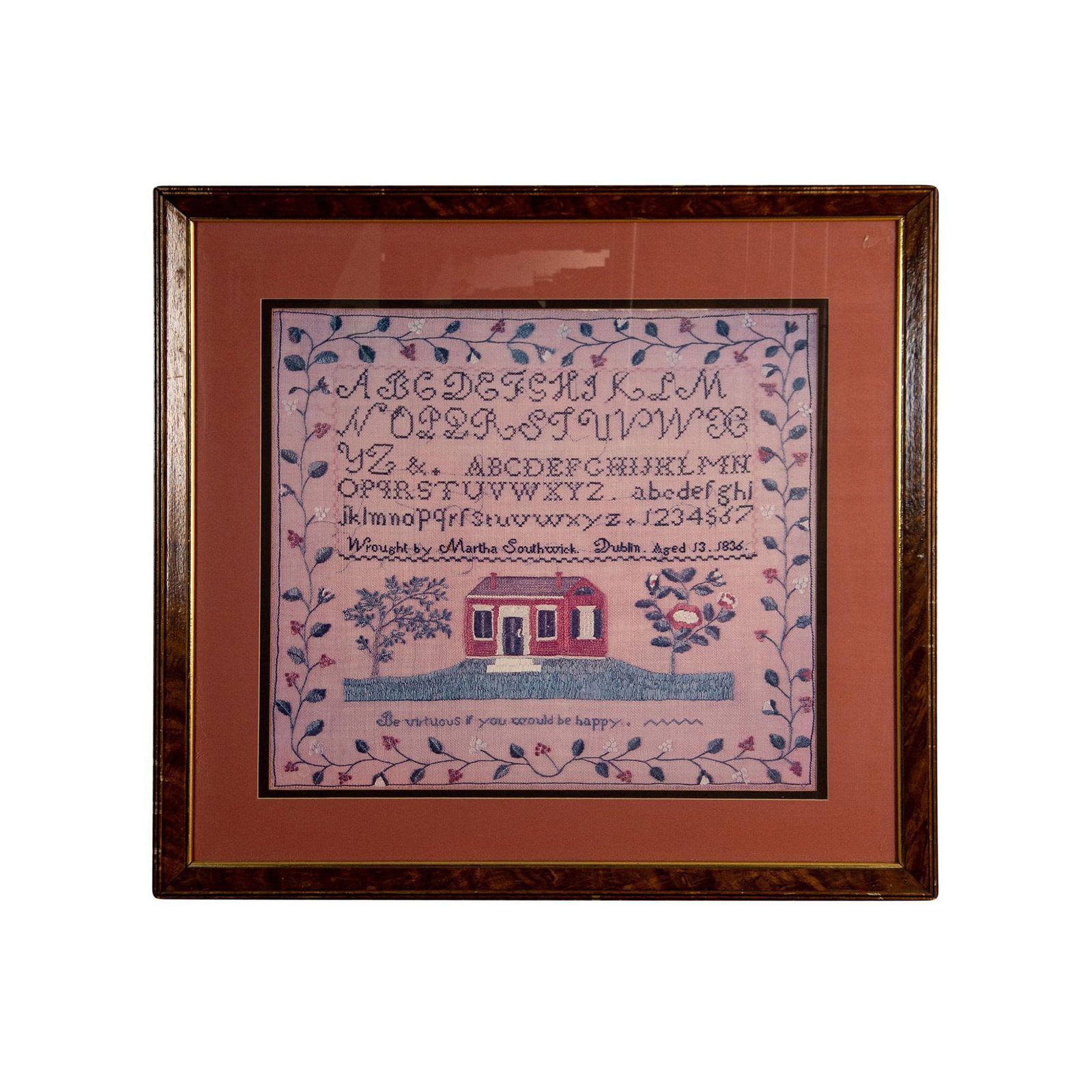 Photo of a 1836 Needlework Pattern by Martha Southwick: Housed in a wooden frame with a pink mat. Depicts an embroidered stitching of a house and the alphabet. Frame size: 27.25"W x 26"H x 1"D. Sight size: 18.75"W x 17.75"H.