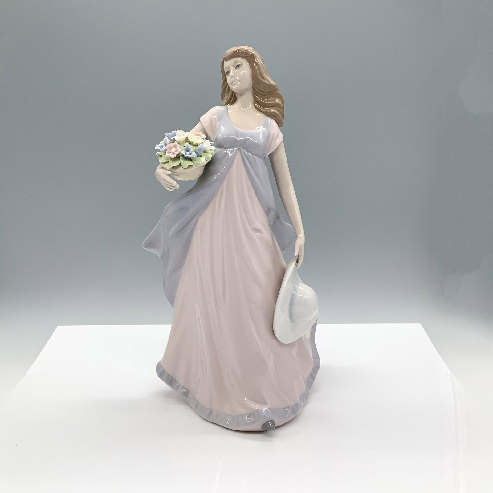 Nadal Porcelain Figurine, Woman with Basket of Flowers: A glossy figure depicting a woman wearing a long flowing dress while holding a basket of flowers in one hand and a wide-brimmed hat in the other. Nadal backstamp. Dimensions: 7"L x 7"W x 13.25"H Manuf