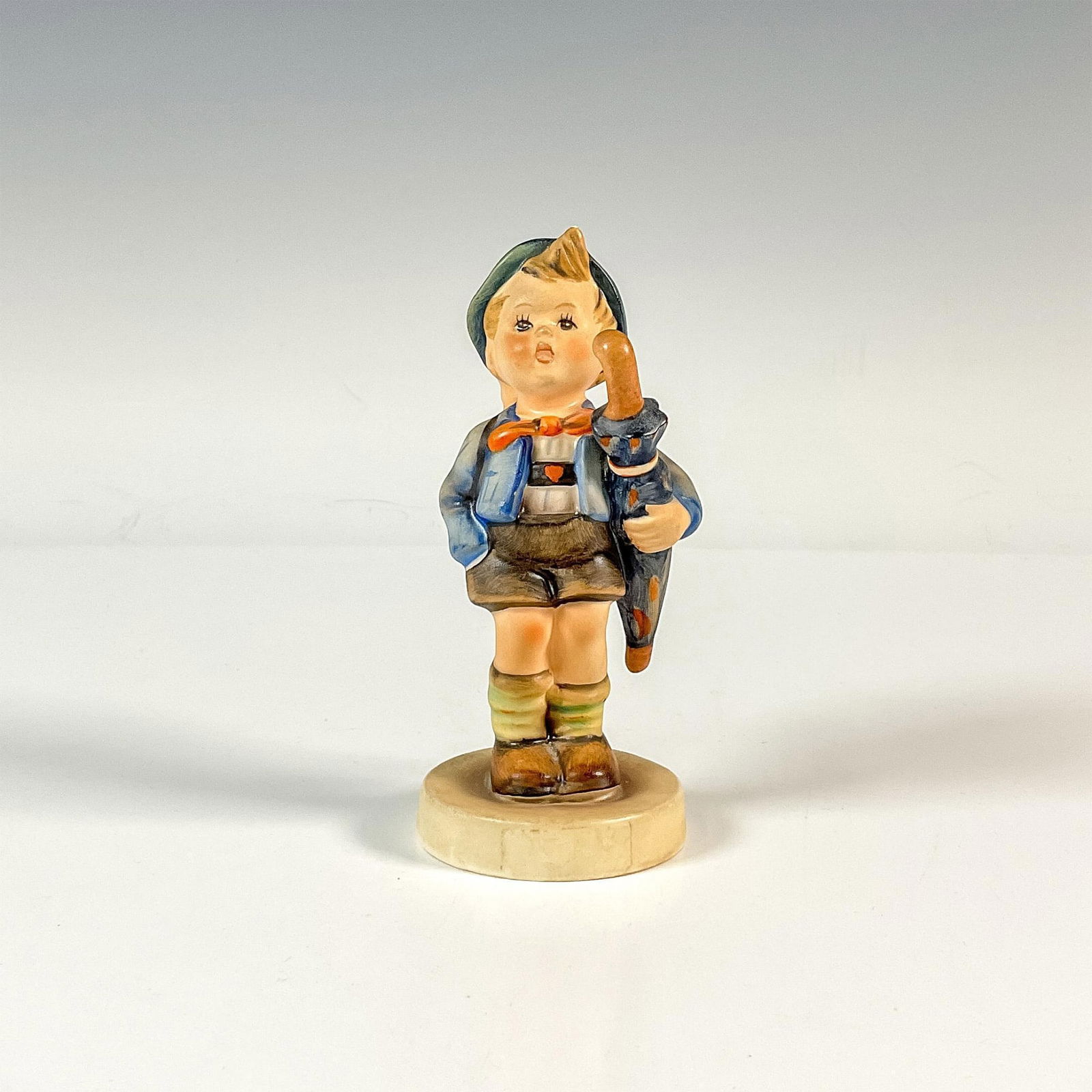 Goebel Hummel Porcelain Figurine, Home From Market: A colorful matte figure of a boy holding an umbrella under his arm and basket with a pig on his back. M.I. Hummel impressed. Goebel backstamp. Issued: 1964-1972 Dimensions: 2"L x 2.5"W x 4.75"H Manufa
