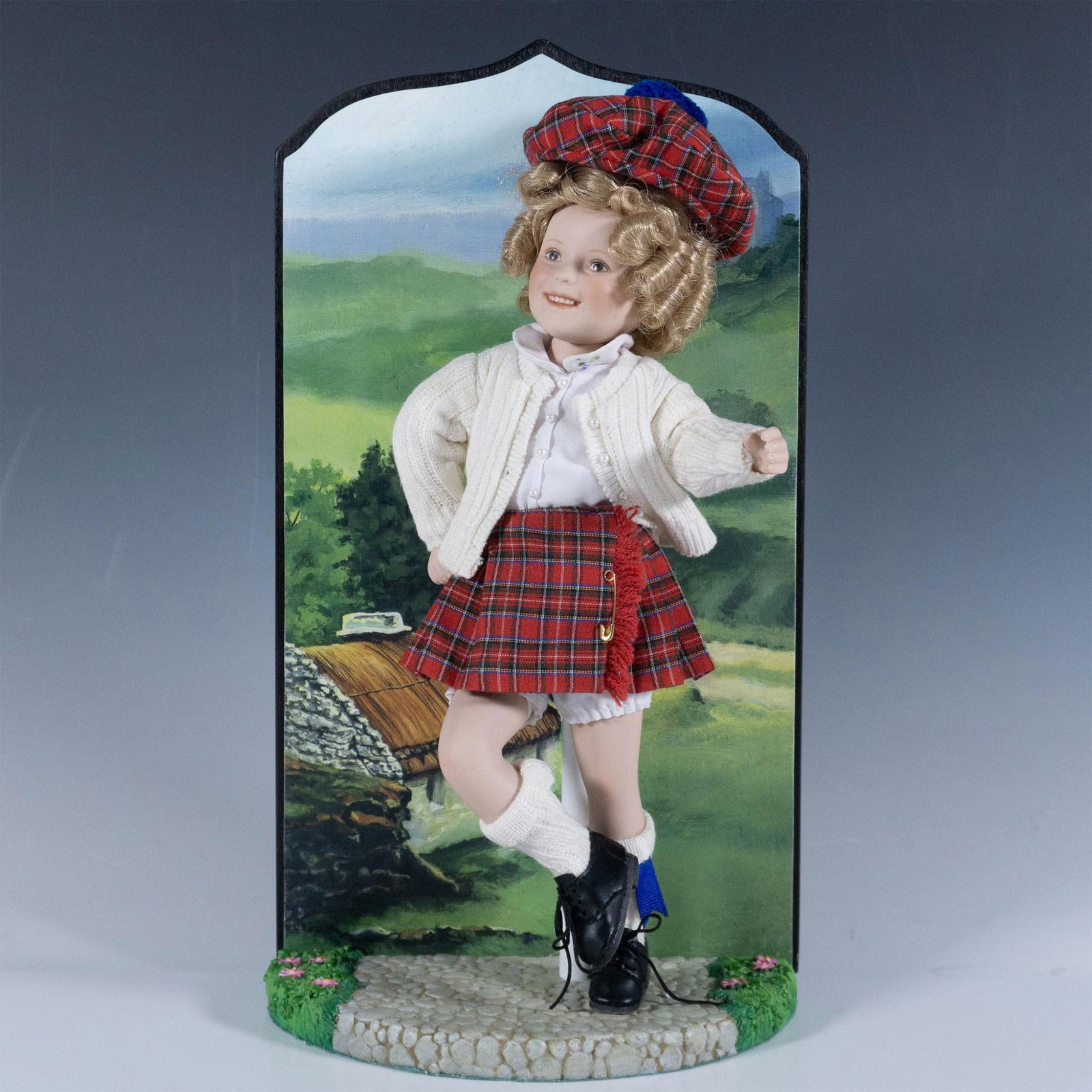 Danbury Mint, Little Lass Shirley Temple Porcelain Doll: Delightful figure of a little girl with curly blonde hair wearing Scottish attire. She stands atop a base and in front of a background. Metal stand included. This item has its original box 9"L x 6.25"