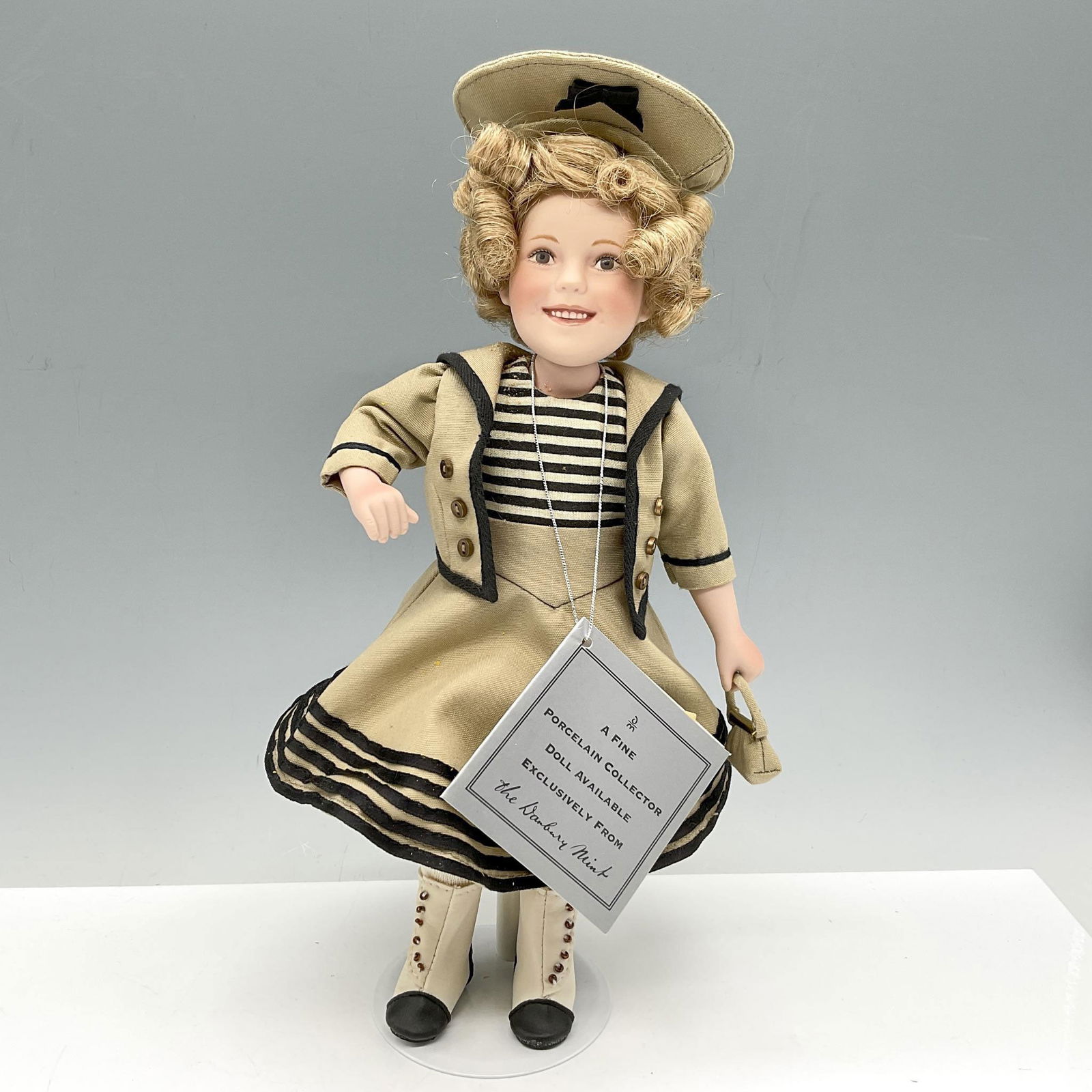 Danbury Mint Porcelain Shirley Temple Movie, Stowaway: A nod to Shirley Temple's movie classics with this hand painted and dressed in hand sewn outfits related to that movie. Styled with matching sailor hat to her khaki suit with black stripes. Metal stan