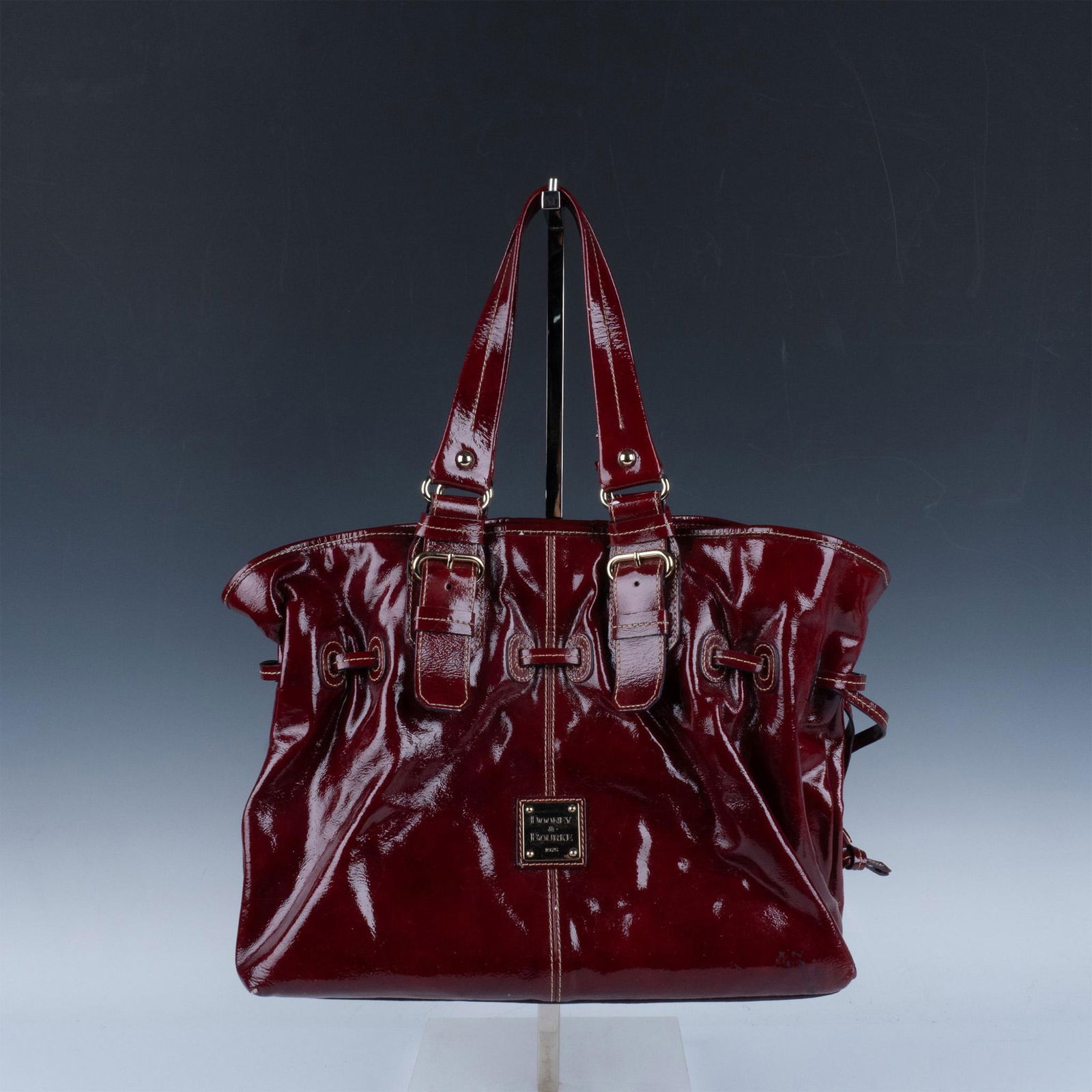 Dooney & Bourke Red Patent Leather Handbag: Vibrant red leather with cinch strap and gold-tone hardware buckle accents. Interior is red striped fabric with center and side zipper pockets plus slip pockets. Dooney & Bourke embossed tag. Issued: