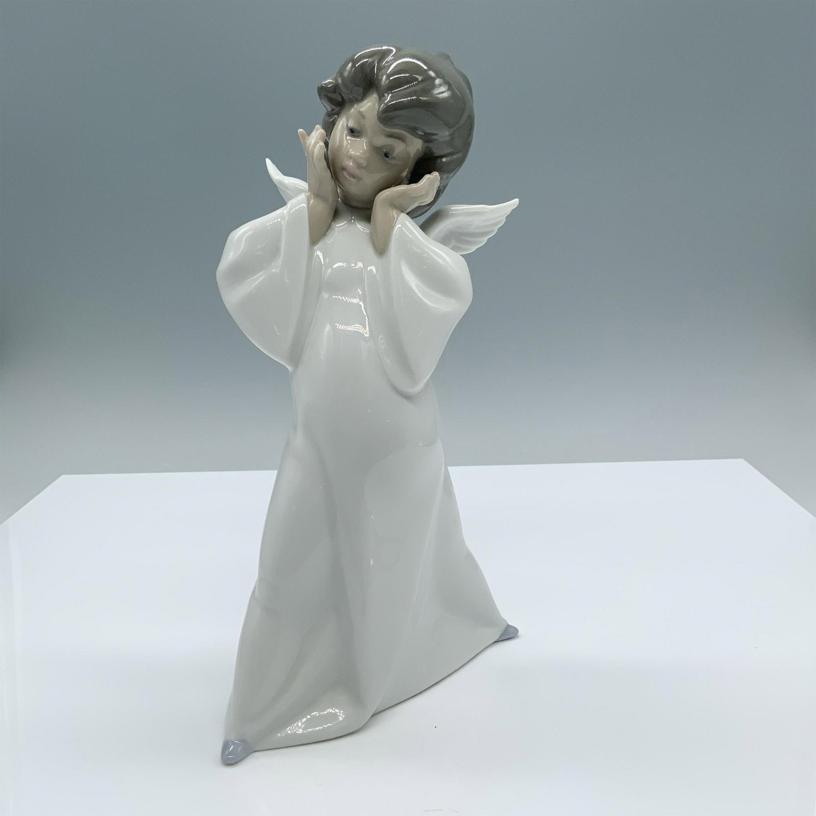 Cherub Puzzled 01004959 - Lladro Porcelain Figurine: Glossy finish. Features a charming angel dressed in a white robe holding his face. Lladro backstamp. Artist: Salvador Debon Issued: 1977 Dimensions: 5.75"L x 4.25"W x 8.75"H Manufacturer: Lladro Coun