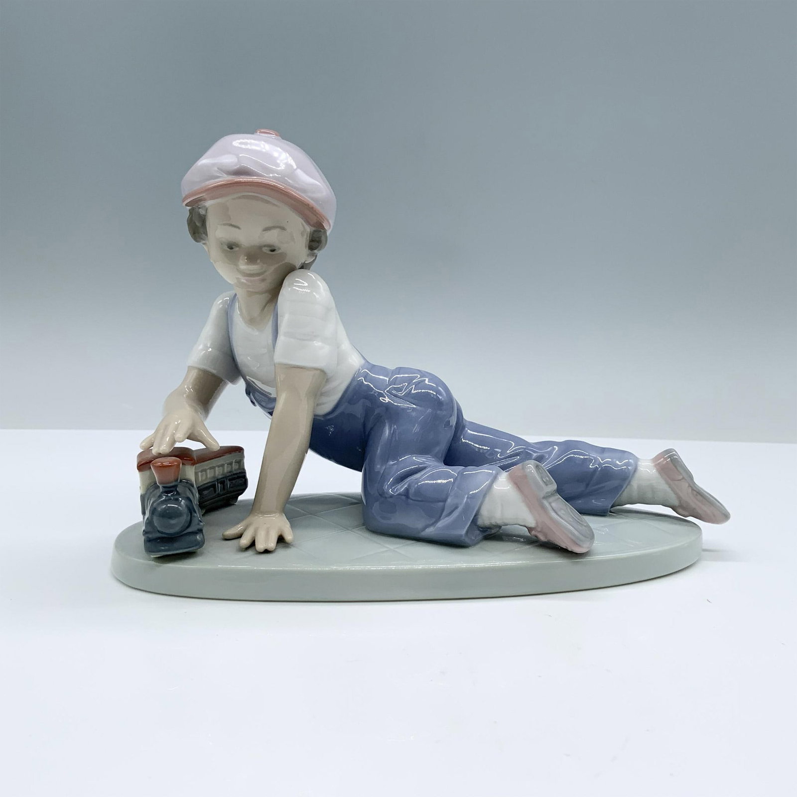 All Aboard 1007619 - Lladro Porcelain Figurine: Little boy playing with a toy train. Glossy finish; Lladro backstamp. Artist: Antonio Ramos Issued: 1992 Dimensions: 7.5"L x 3.25"W x 5"H Manufacturer: Lladro Country of Origin: Spain