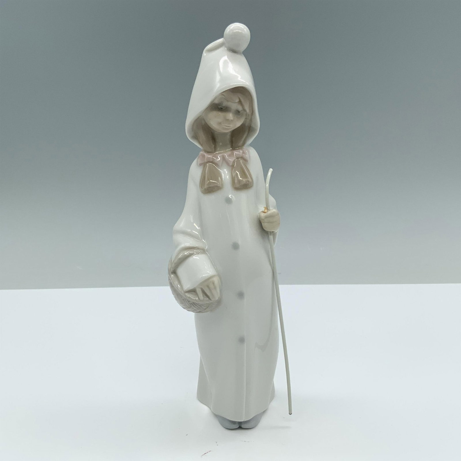 Girl with Basket 01004678 - Lladro Porcelain Figurine: Glossy finish. Features a girl standing with a white hooded robe, a cane, and a basket. Lladro backstamp. Artist: Juan Huerta Issued: 1969 - 2001 Dimensions: 2"L x 2.25"W x 8.75"H Manufacturer: Lladro