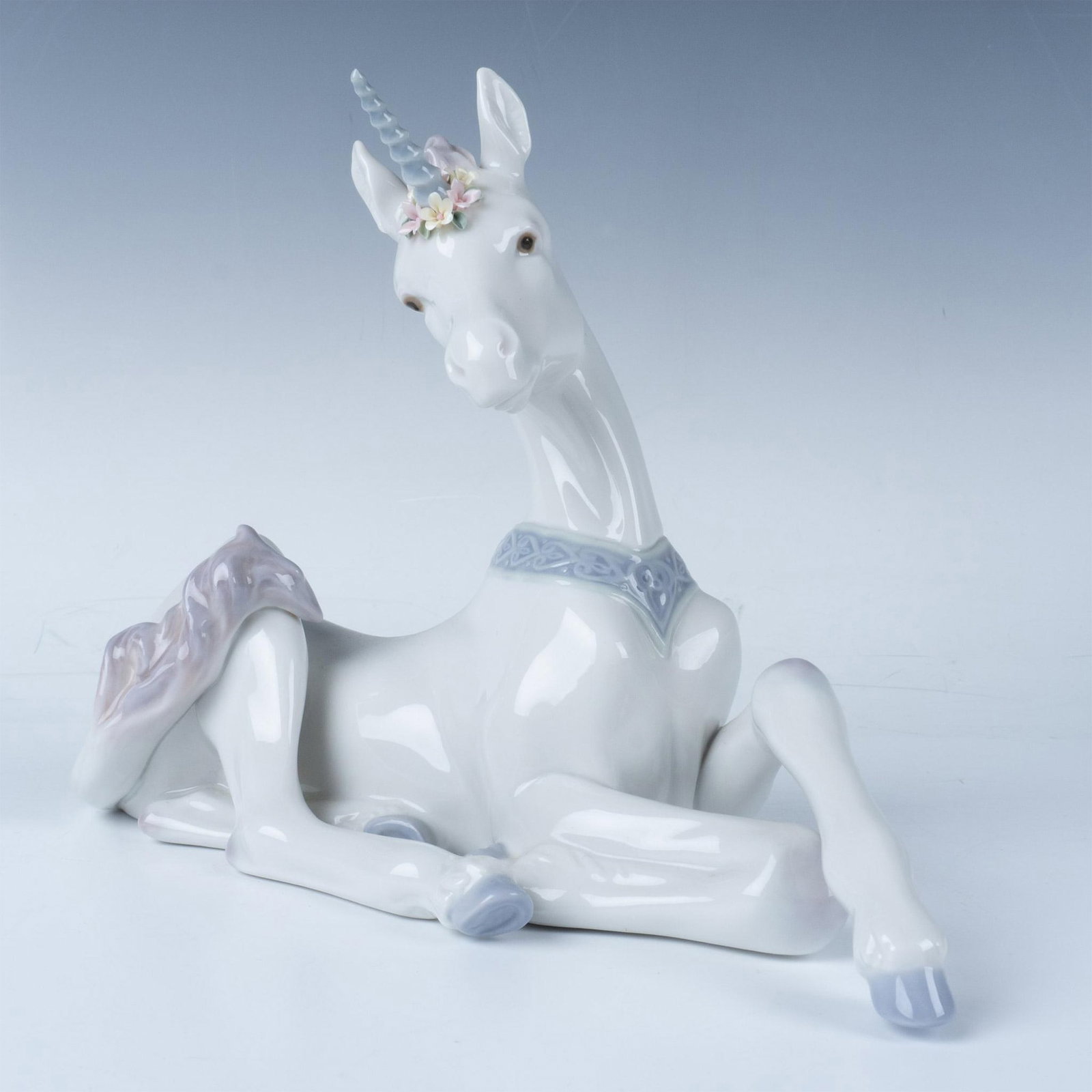 Little Unicorn 1005826 - Lladro Porcelain Figurine: Glossy finish. Features a white unicorn with purple accents, laying down on the floor with flowers around its horn. Lladro backstamp. This item has its original box. Box measures 10.25"L x 12.5"W x 6"