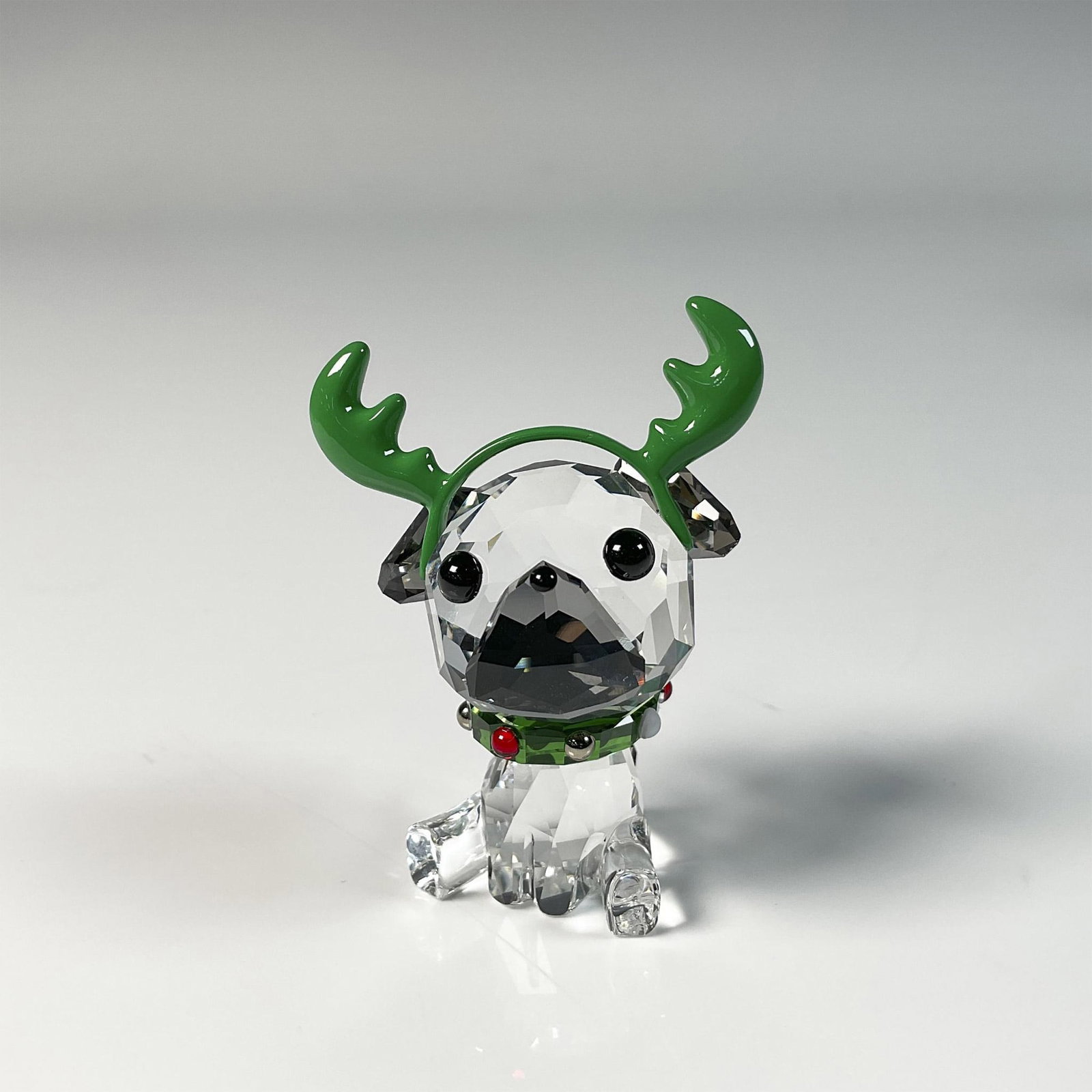 Swarovski Crystal Figurine, Holiday Cheers Pug: Adorable figure pug, crafted from clear crystal and adorned with a Santa headband and a necklace featuring a green accent, capturing the joyful spirit of Christmas. Swarovski swan mark. This item incl
