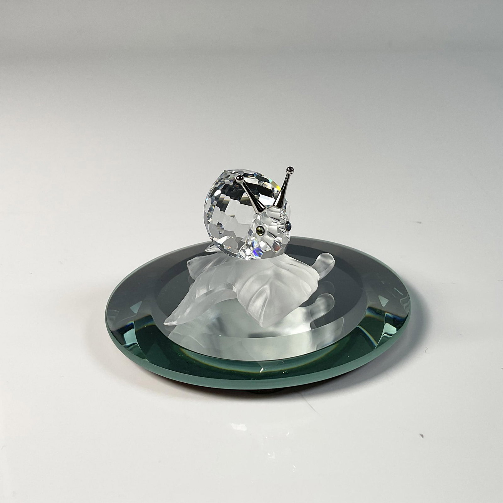 Swarovski Crystal Figurine and Base, Snail: Adorable snail figure, crafted from clear crystal featuring frosted silver accent on a vine leaf, showing an intricate design. Swarovski swan mark. This item includes a mirror base: 0.25"H x 3" dia. o