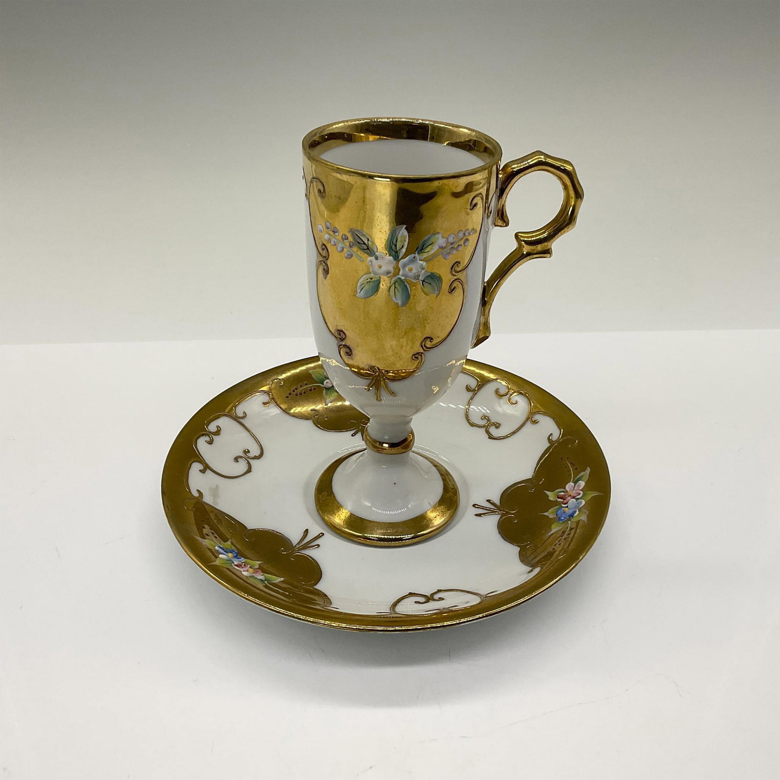 2pc Royal Carlton Painted Glass Coffee Cup and Saucer: Beautiful tall coffee cup and matching saucer adorned with enameled flowers and embossed gold motifs over fine white bone china. Royal Carlton backstamp. Cup is 3"L x 2.25"W x 4.75"H. Saucer is 5.75"W