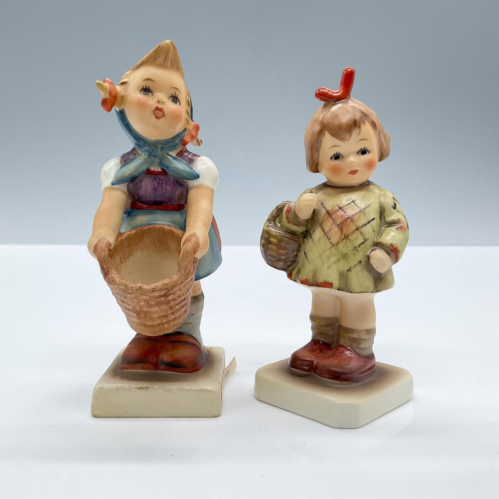 2pc Goebel Hummel Figurines, Little Helper & Bought You Gift: This adorable duo features two little girls with baskets, includes: Little Helper 73 and I Bought You a Gift. Tallest piece dimensions: 1.5''L x 2.25''W x 4.25''H. Goebel Hummel backstamp. Issued: 20t