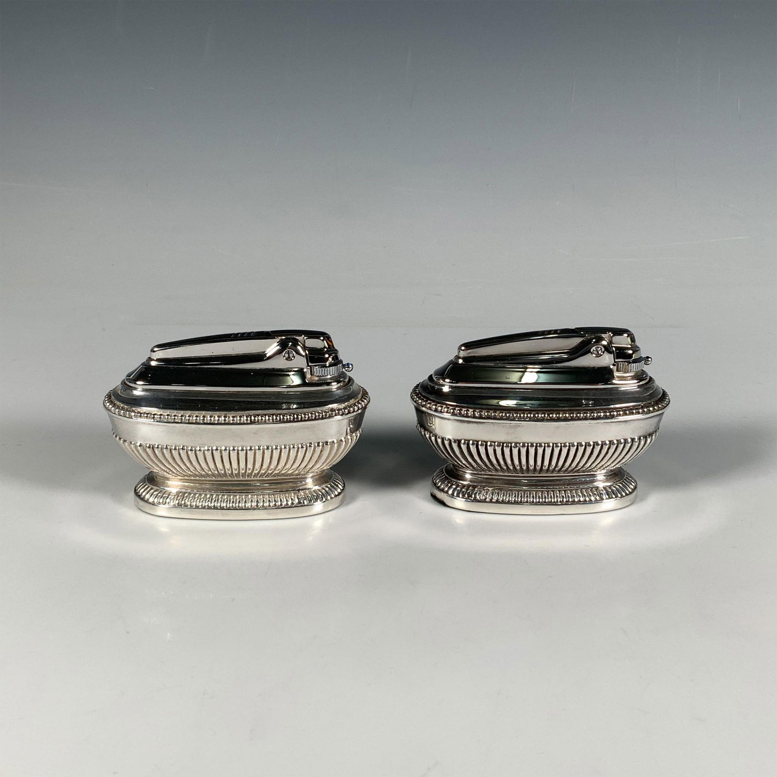 2pc Ronson Queen Anne Varaflame Silverplate Table Lighters: This opulent set includes two vintage Art Deco silver-plated table lighters, each featuring an ornate ribbed pattern and adorned with a delicate soft blue gemstone, inspired by the design of the Queen