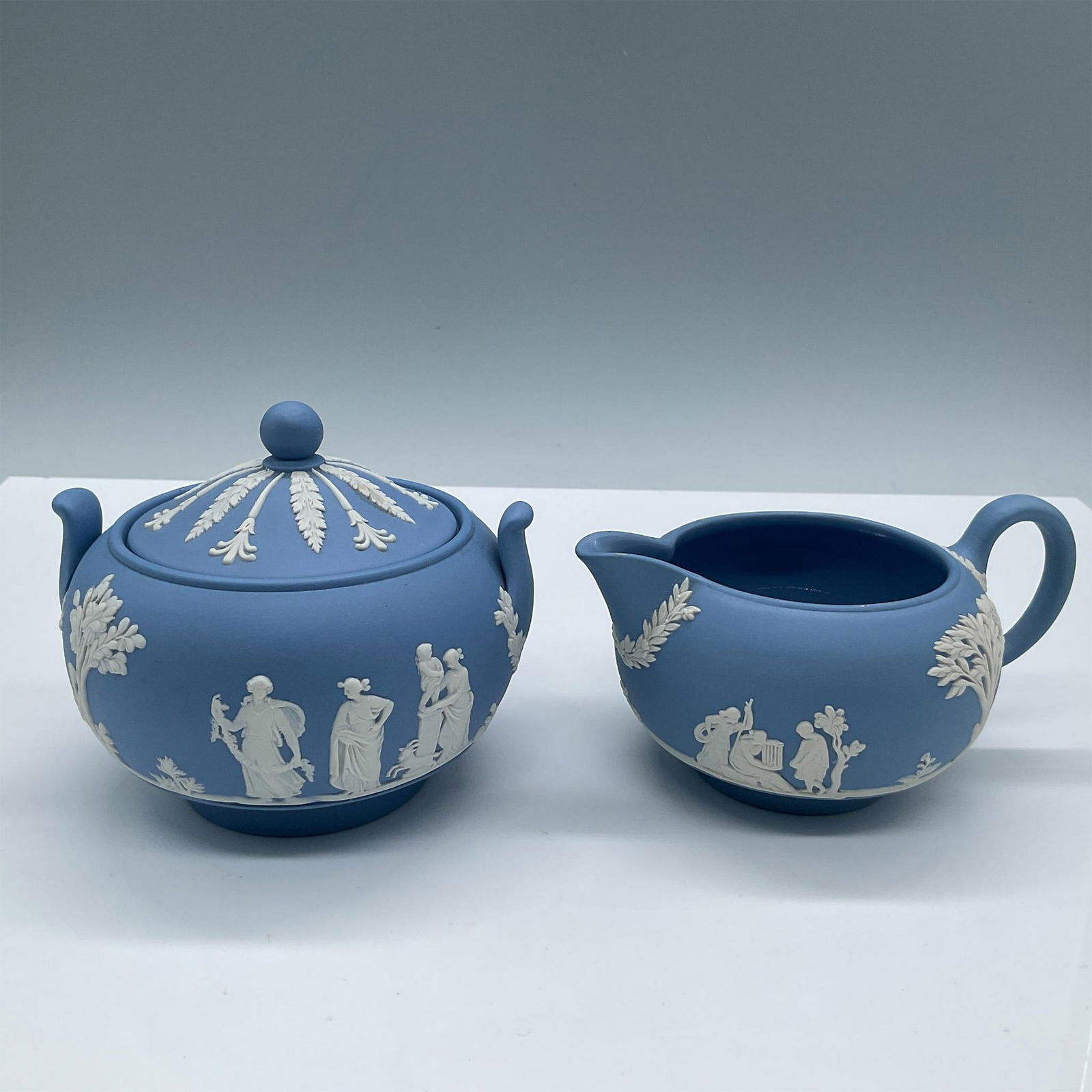 2pc Wedgwood Pale Blue Jasperware Creamer & Covered Sugar: Beautiful duo of pale blue English pottery. Includes a lidded sugar bowl with two little handles, alongside a creamer jug. In white cameo reliefs: Sacrifice figures. Wedgwood impressed marks on bases.