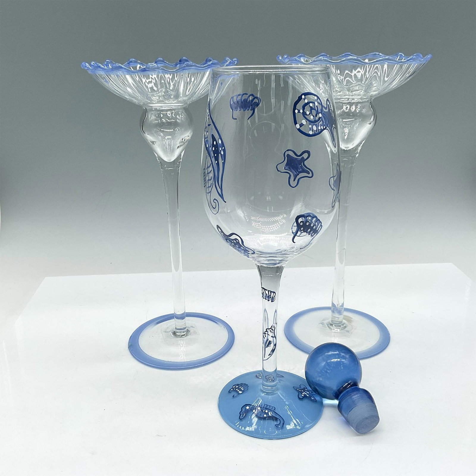 4pc Blue Tableware Collection, Candle Holders + more: Assortment of clear glass with blue accents. Lot includes round blue stopper 2.5"H, tall wine glass designed with blue seashell accents 9"H and 2 tall clear holders with blue ruffled edge and base 9"H