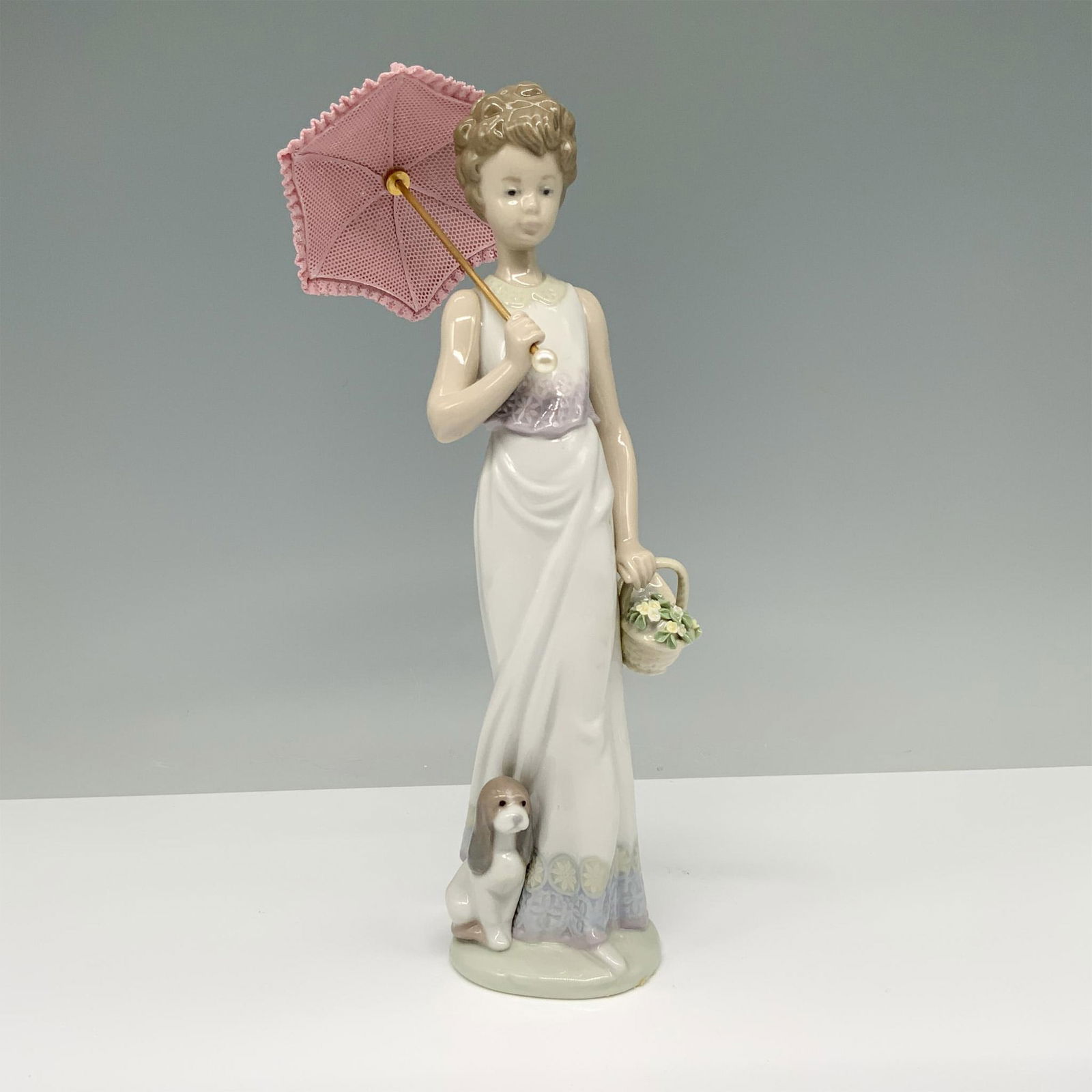Garden Classic 01007617 - Lladro Porcelain Figurine: Glossy finish, depicts a woman in a white sun dress carrying a basket of flowers alongside her dog while holding a parasol. Lladro backstamp. This item has its original box. Box measures: 13"L x 7.5"W