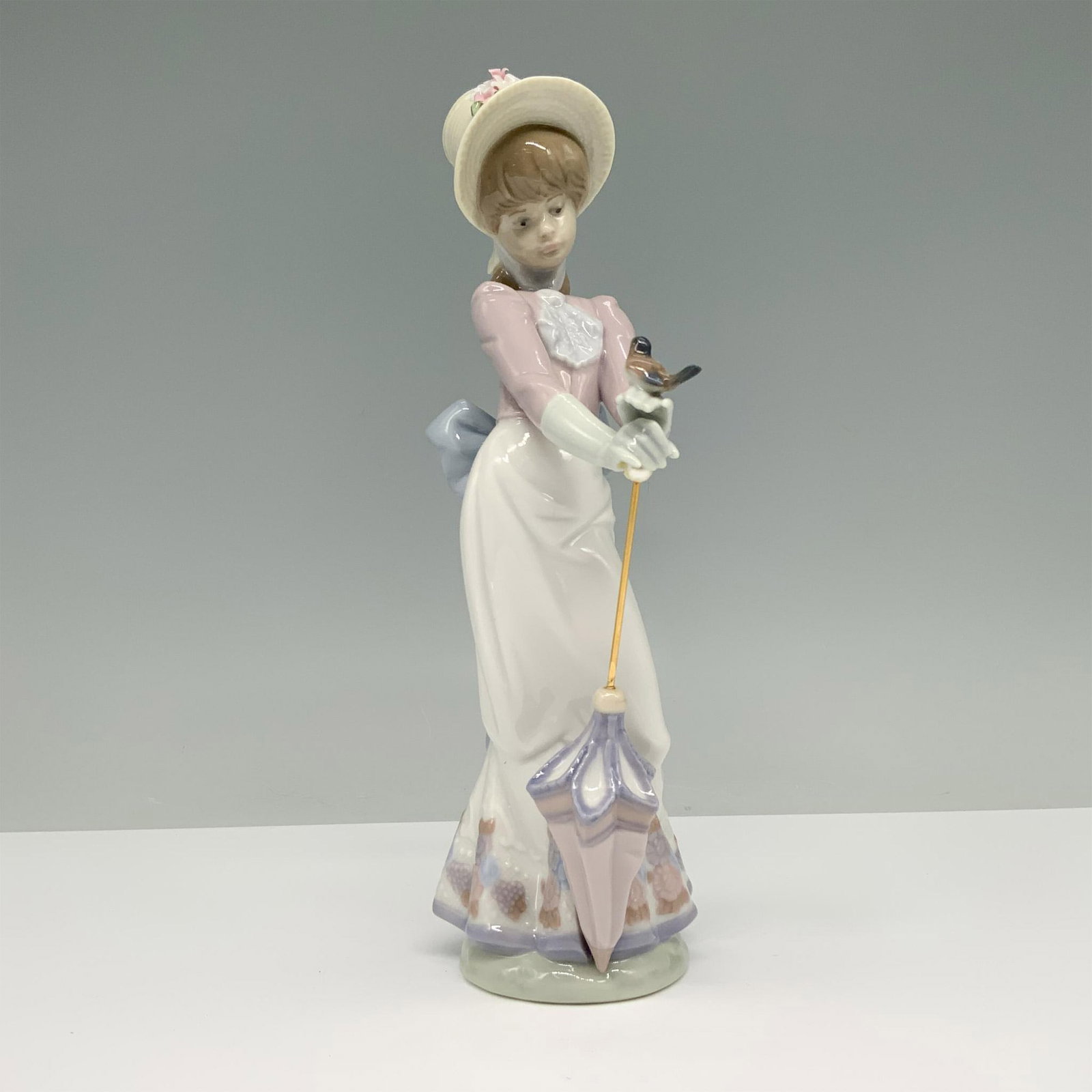 Garden Song 01007619 - Lladro Porcelain Figurine: Glossy finish porcelain figurine depicting a woman in Victorian-style attire holding an umbrella and a small bird in her hand. Lladro backstamp. This item has its original box. Box measures: 11"L x 5.