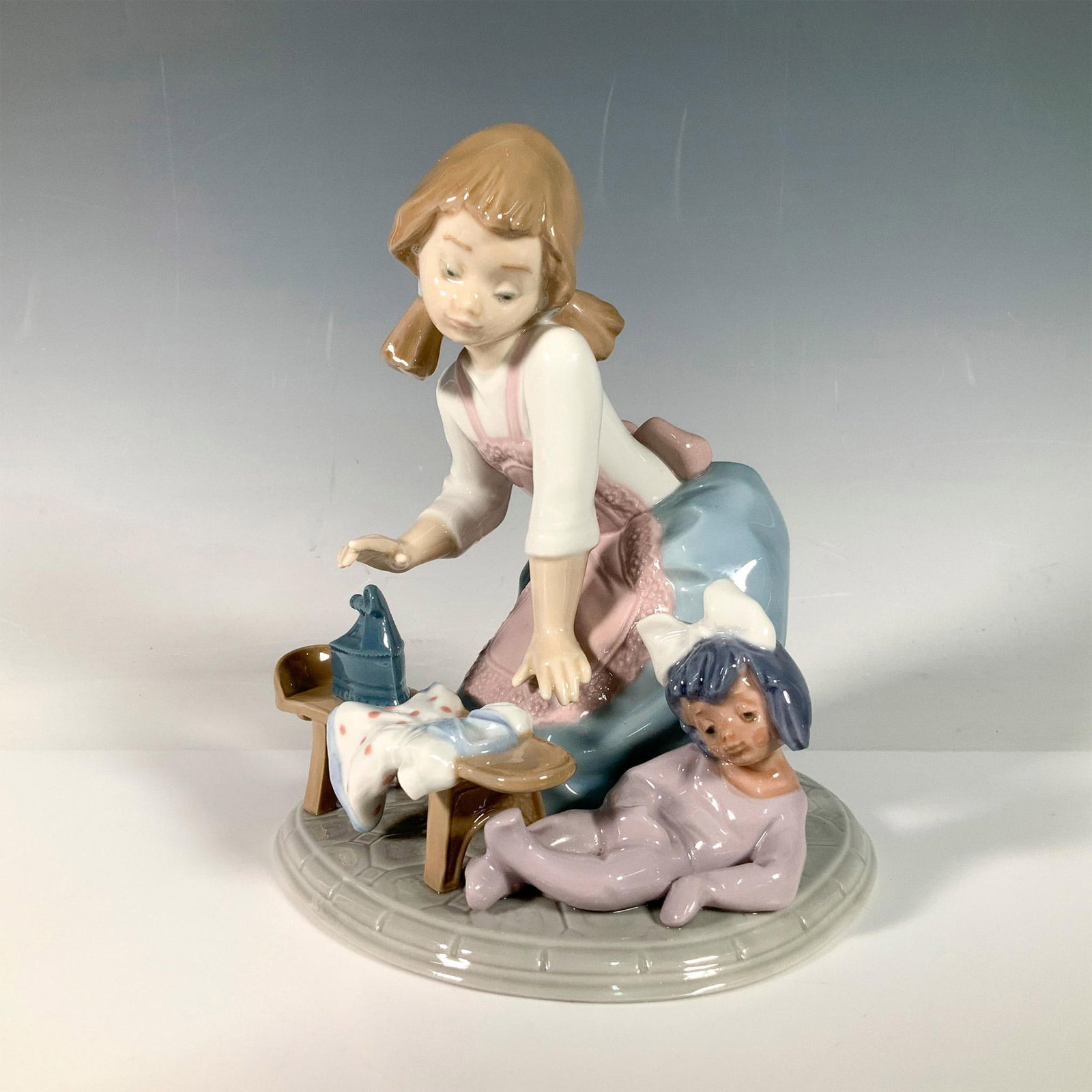 My Chores 1005782 - Lladro Porcelain Figurine: My chores features a young girl with brown pony tails dressed in a pink and blue apron using an iron on her younger sister's dress. Lladro backstamp. Artist: Francisco Polope Issued: 1991-1995 Dimensi