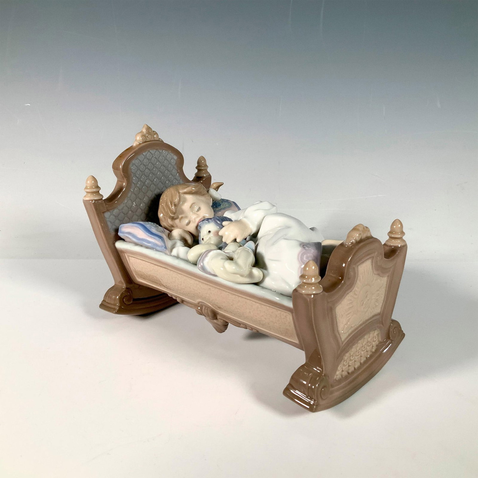 Rock A Bye Baby 1005717 - Lladro Porcelain Figurine: Glossy porcelain figure of a young girl sleeping in a cradle holding her doll. Lladro backstamp. Artist: Francisco Polope Issued: 1990-1999 Dimensions: 6.5"L x 4"W x 4.5"H Manufacturer: Lladro Countr