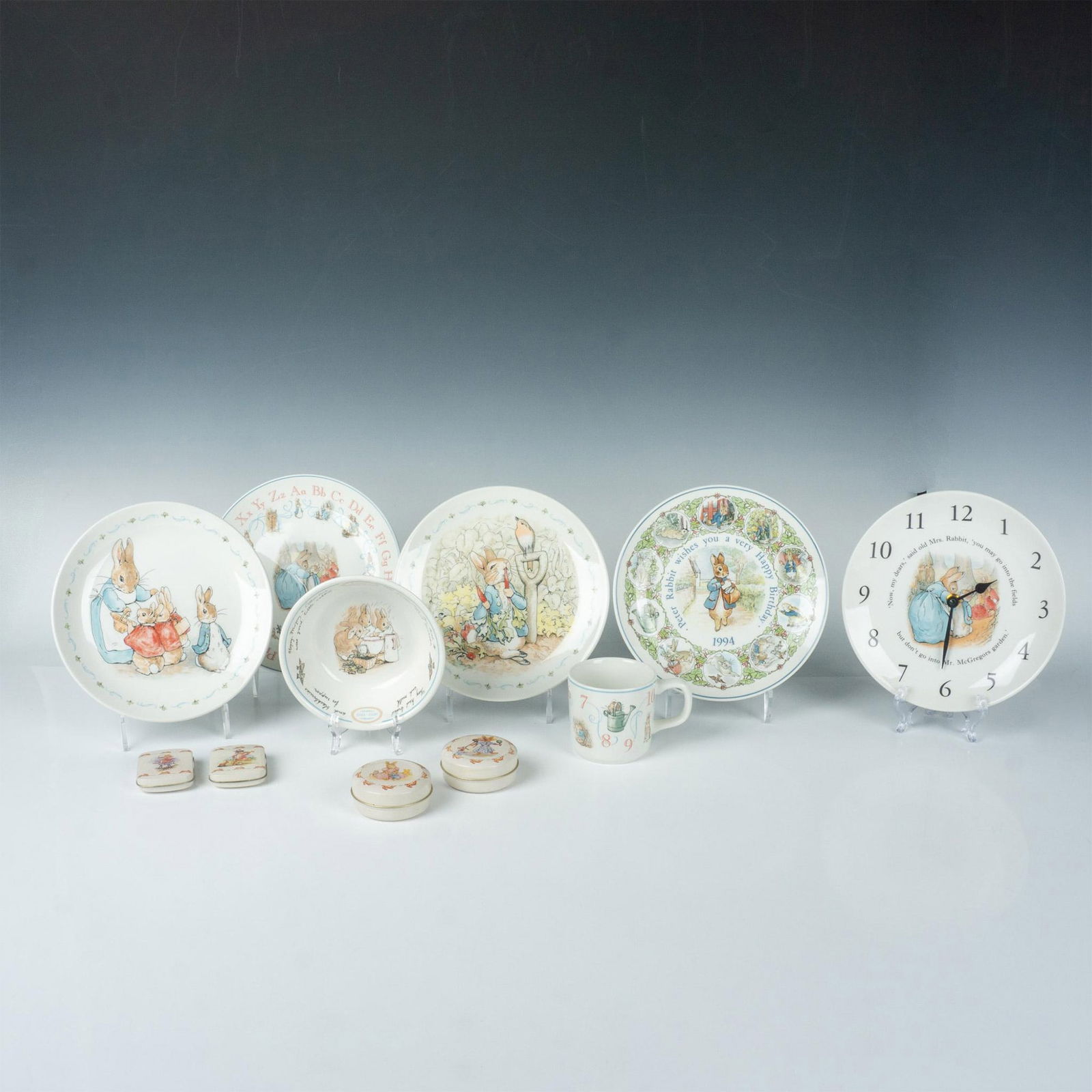 11pc Wedgwood Peter Rabbit Tableware, Clock + Treasure Tins: Cheerful group of decorative Bunnykins items. Wedgewood bone china tableware and clock, stamped Wedgewood. Four luncheon plates 0.75"H x 8" dia. Bowl 1.75"H x 5.75" dia. Small mug 4.25"L x 3"W x 2.75"