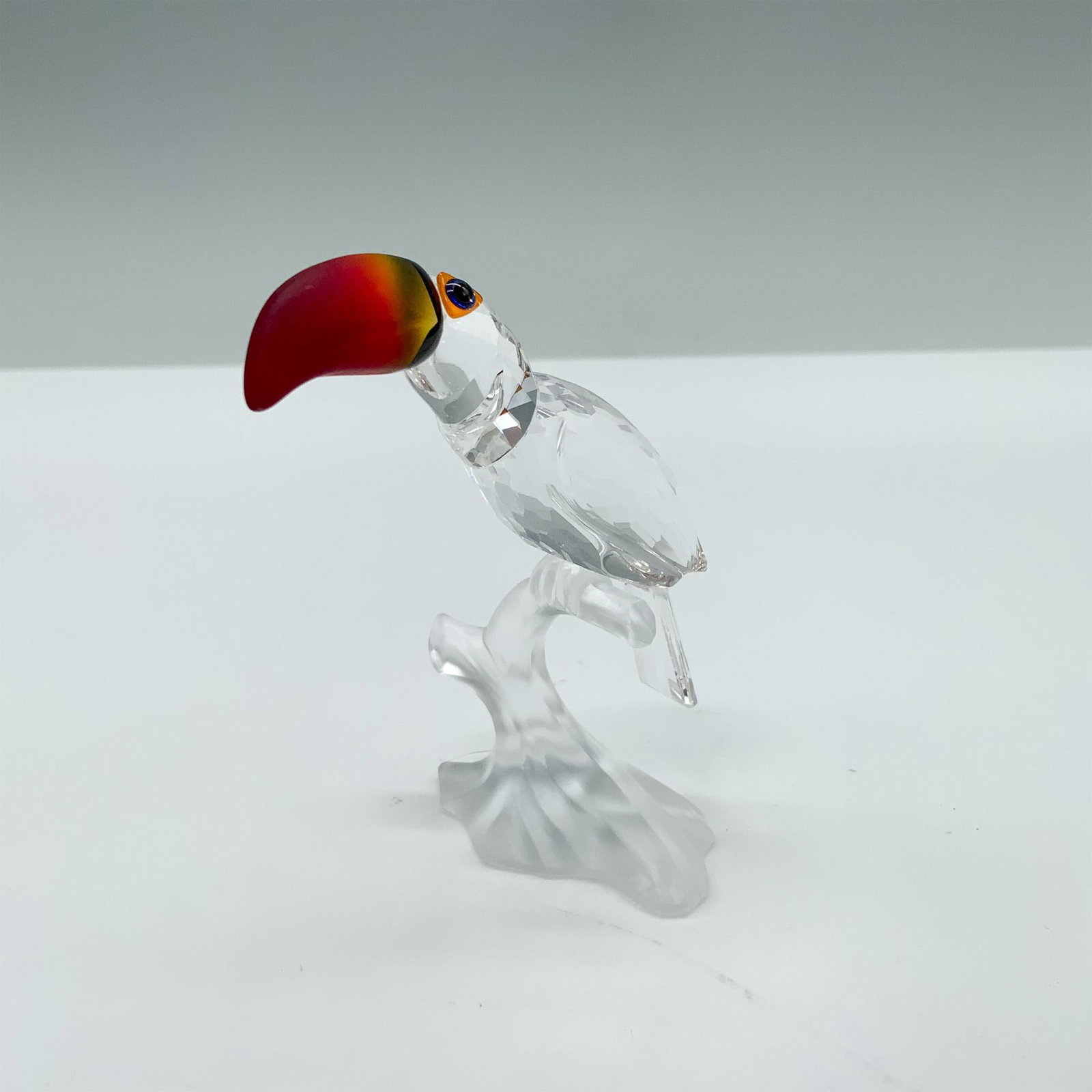 Swarovski Crystal Figurine, Toucan: This crystal toucan was made from clear crystal and sits on a frosted branch. His beak is made from fire opal crystal. The toucan was part of the Feathered Beauties collection. Swarovski swan mark. Th