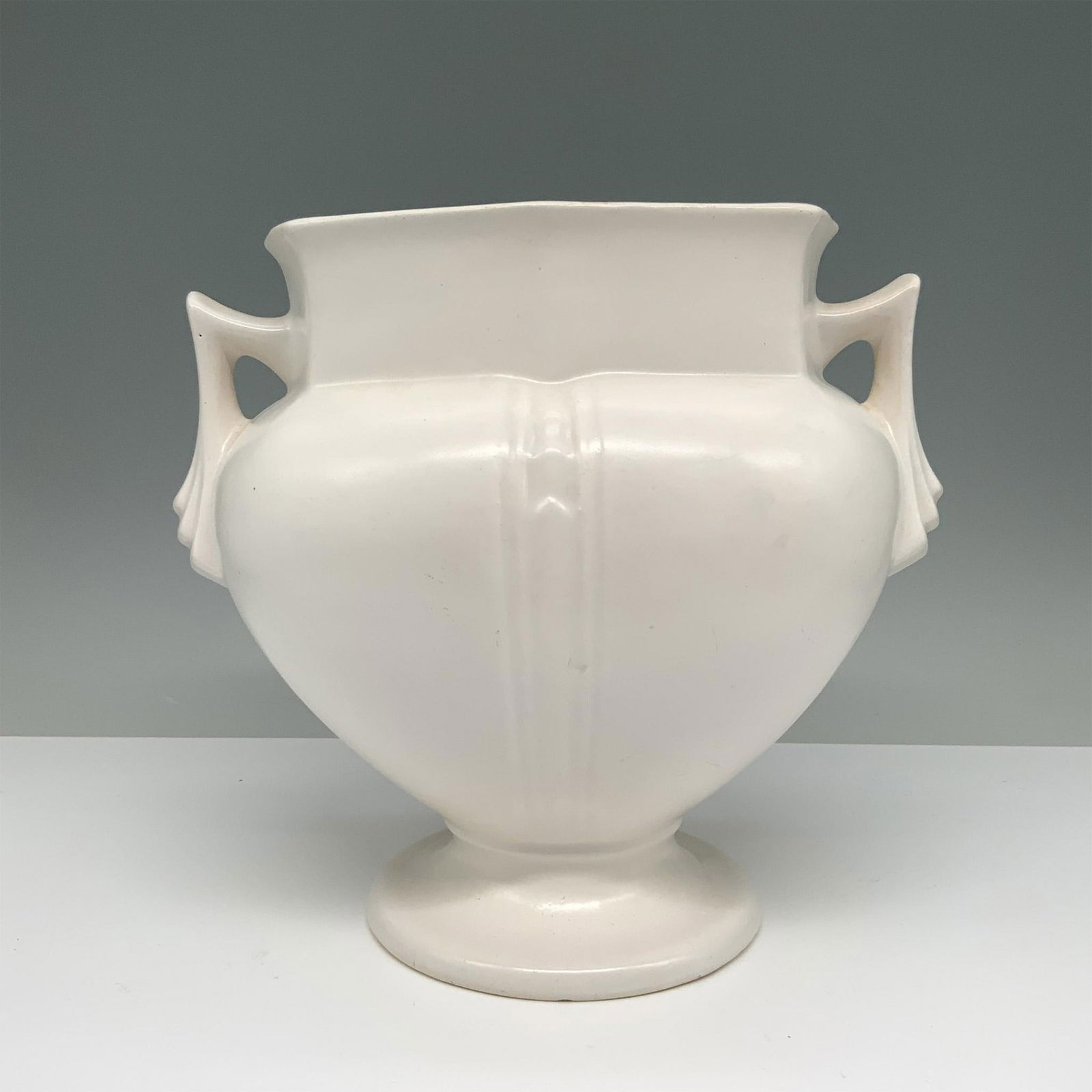 Amphora Pedestal Vase: Refined vintage ceramic vase featuring a creamy white glaze, adorned with dual looped handles and a gently rounded body. Set on a pedestal base, embodies classic elegance. Dimensions: 8.5"L x 5"W x 7.