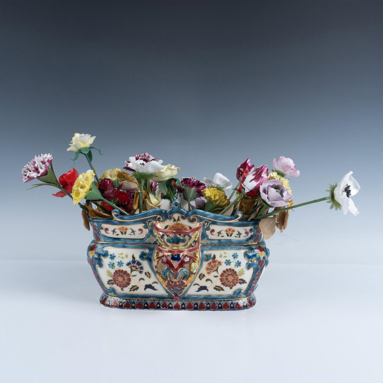 Vintage Porcelain Jardiniere and Flowers: A lavish jardiniere embellished with a painted floral motif and gilded gold. The Jardiniere is filled with an array of 28 porcelain flowers on plastic stems with velveteen leaves. Jardiniere measures 