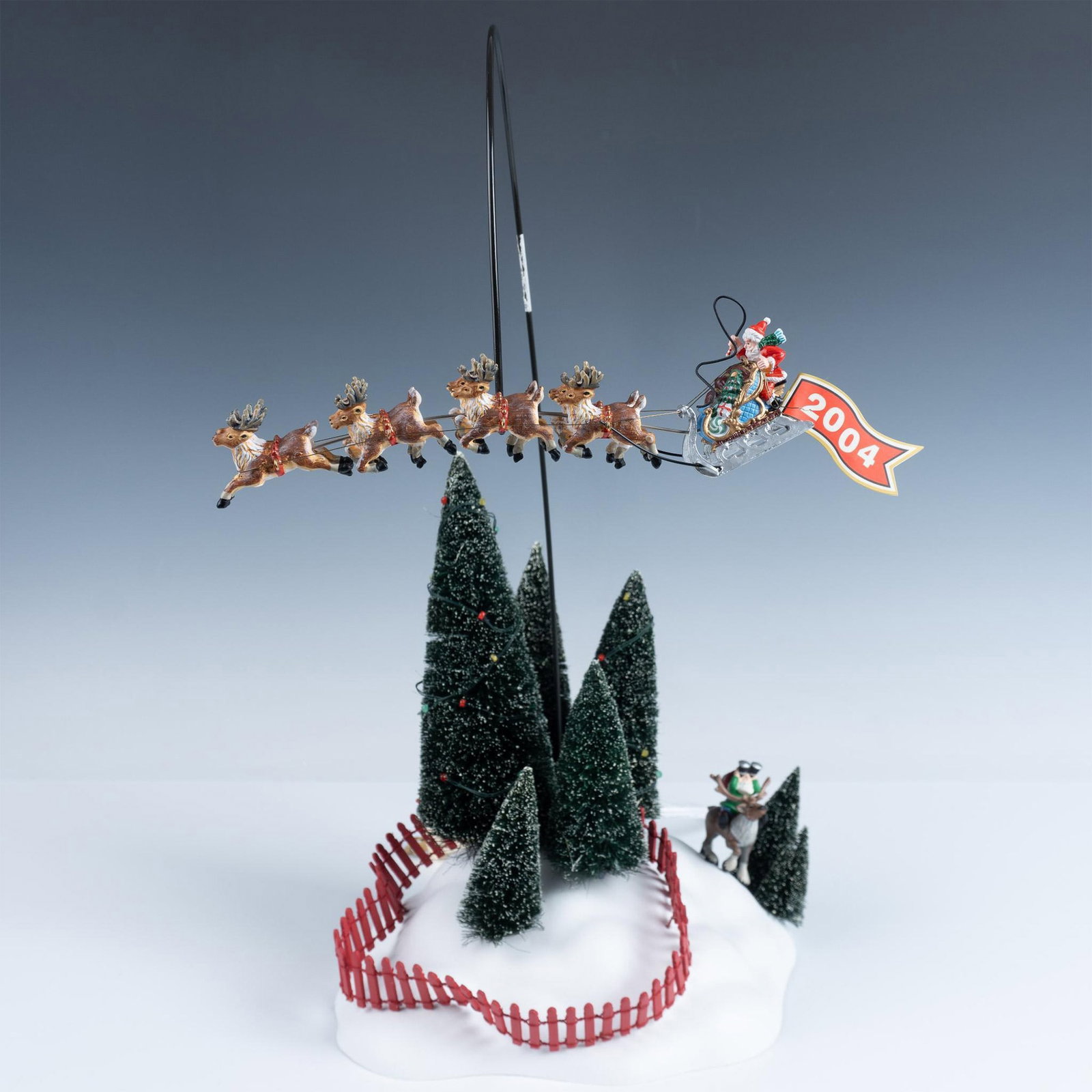 Department 56 Figurine, Santa's On His Way: A snowy hillside, framed by decorated pine trees and a red picket fence, features an elf on a reindeer peering through binoculars at Santa's sleigh as it flies away. Department 56 backstamp. This item