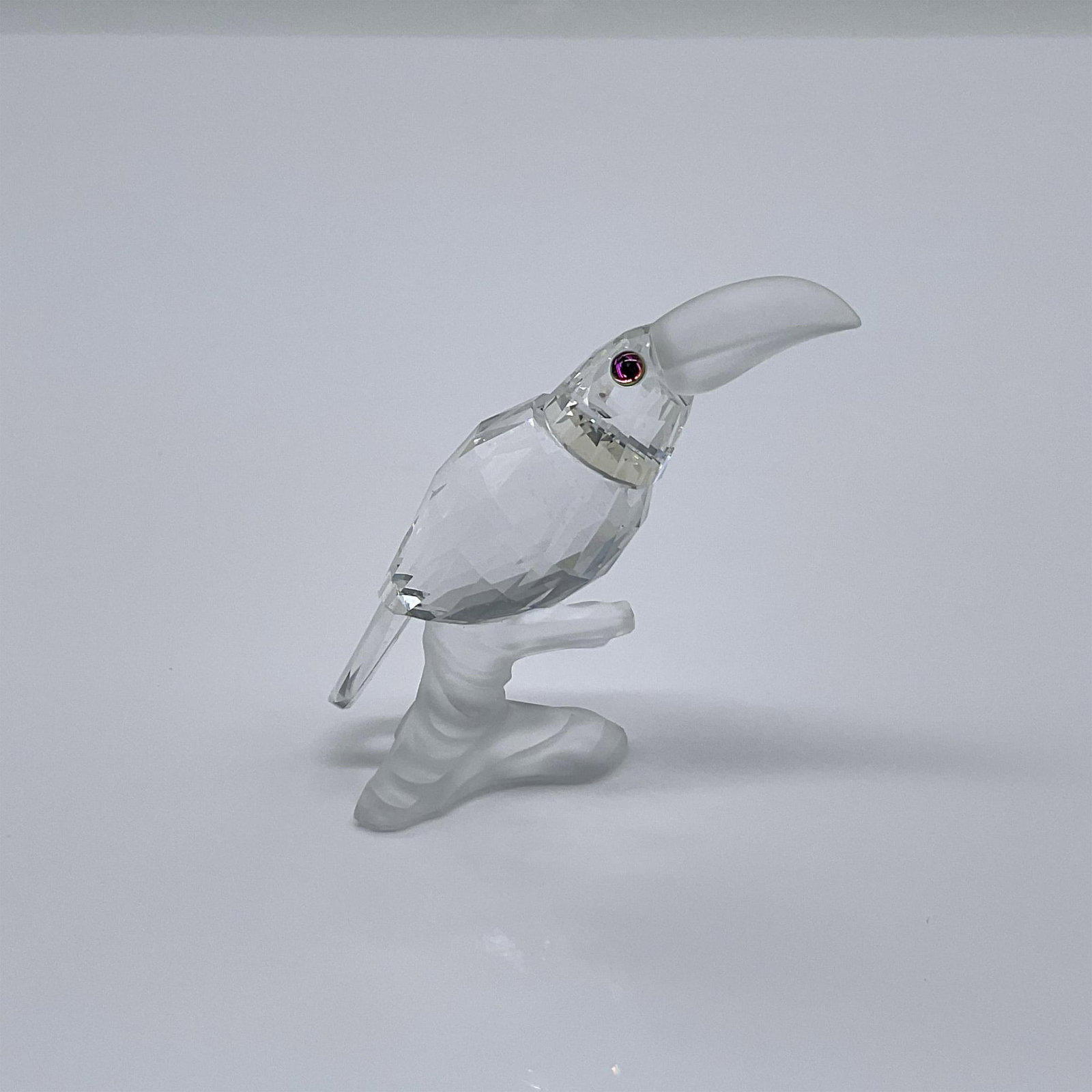 Swarovski Crystal Figurine, Toucan On Branch: From the collection, Up In The Tree. A clear crystal bird with a frosted beak, resting on a frosted branch base. Swarovski swan mark. This item has its original box: 3.25"W x 5"H. Artist: Michael Stam