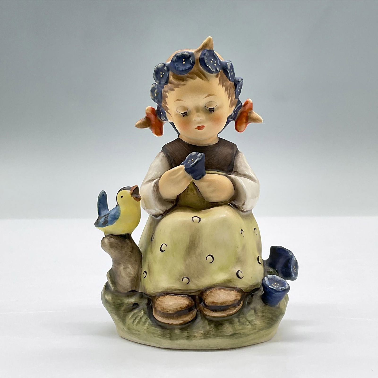 Goebel Hummel Figurine, Botanist: Features a precious porcelain girl with pig tails and a blue flower crown, holding a blue flower while a small bird chirps beside her. Marked Goebel 351. Issued: 1972 Dimensions: 3"L x 2.5"W x 4.5"H M