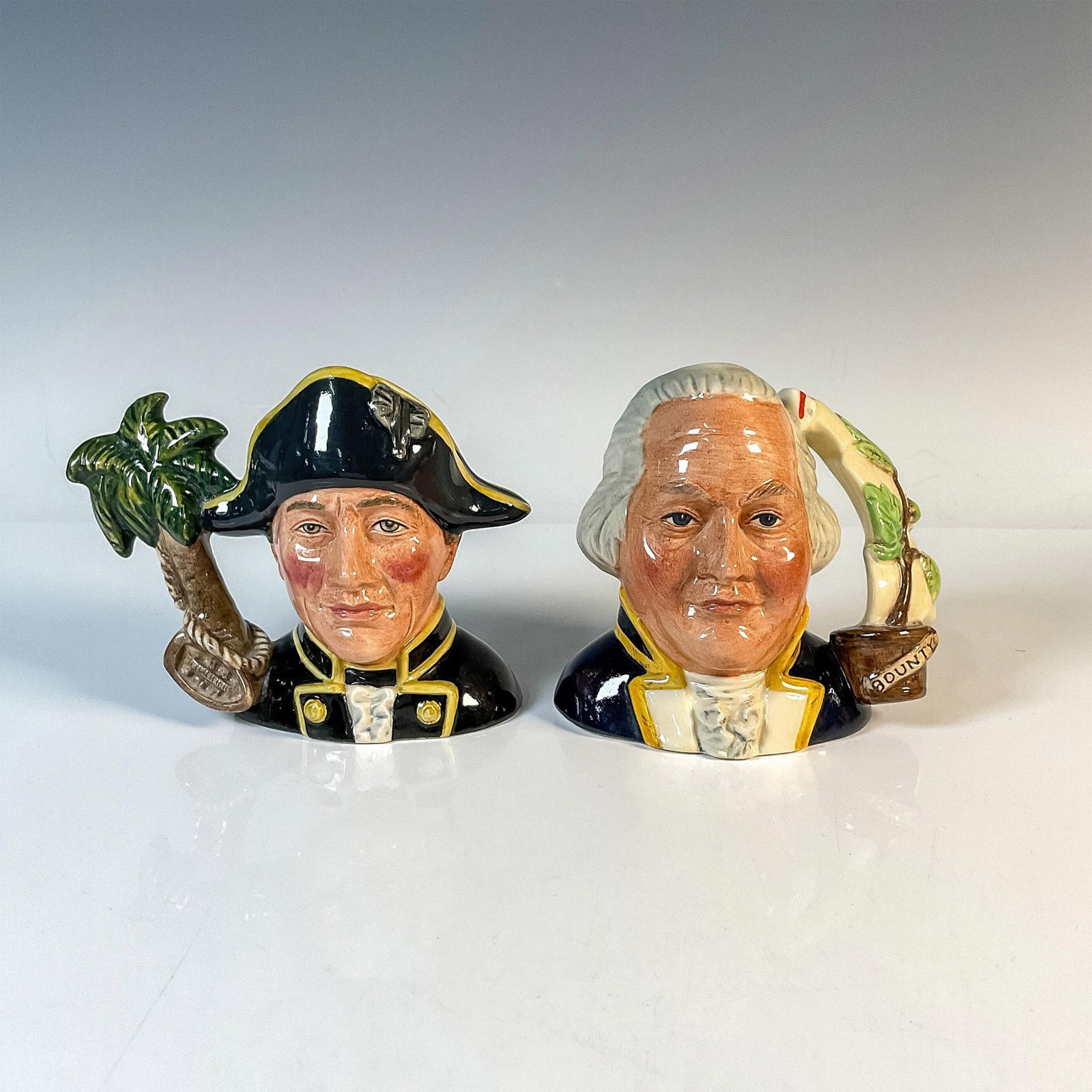 Capt. Bligh and Fletcher Christian D7074 & D7075 - Small - Royal Doulton Character Jug: Set includes: Captain Bligh (black, white, yellow, and brown) and Fletcher Christian (dark blue, white, grey, and yellow). Royal Doulton backstamp. Artist: Robert Tabbenor Issued: 1997 Dimensions: 4.5