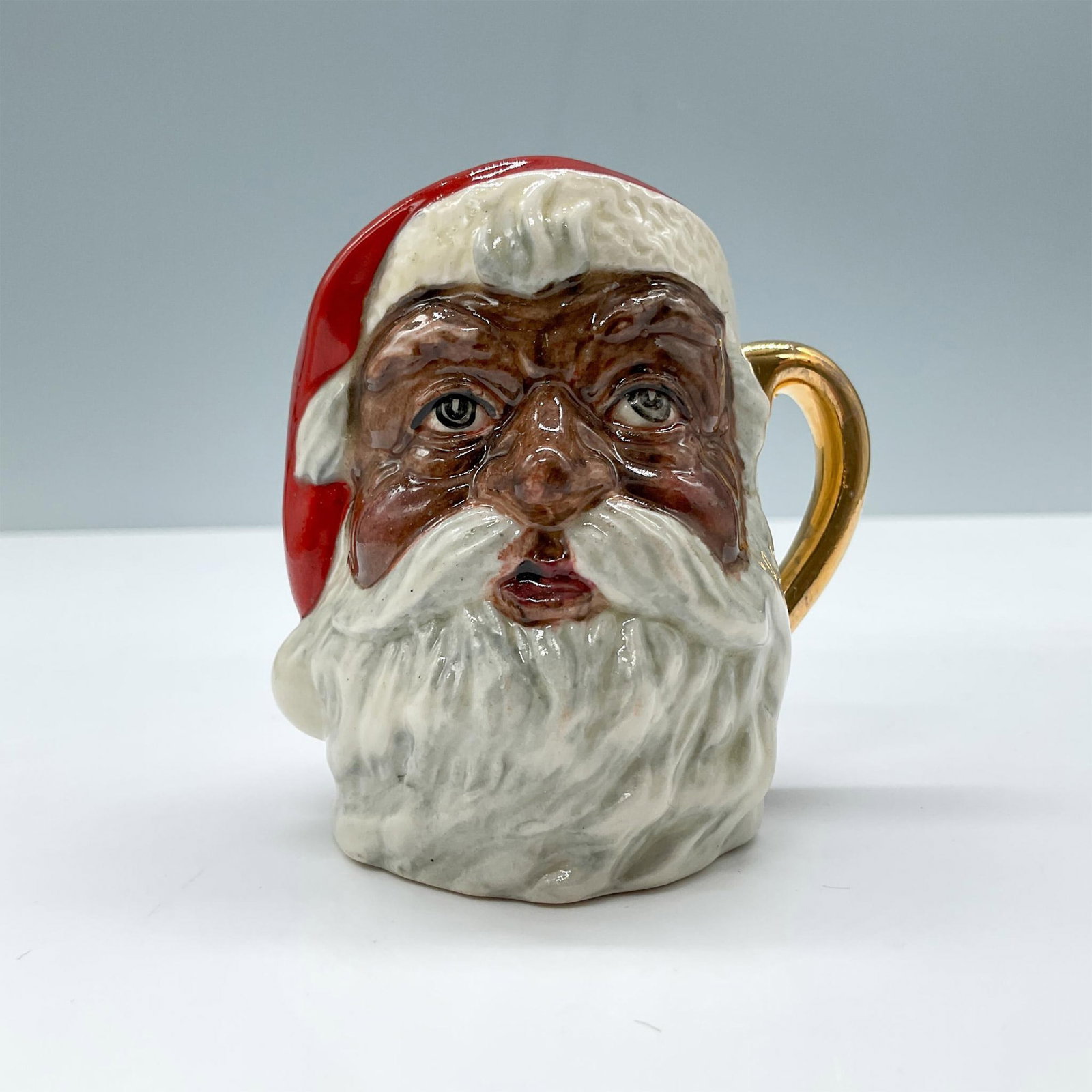 Santa Claus D6706 (Colorway) - Mini - Royal Doulton Character Jug: Figural jug of a Black Santa Claus in red and white. A plain gilt painted handle. Royal Doulton backstamp. Artist: Michael Abberley Issued: c. 1984-1991 Dimensions: 2.75"L x 2.25"W x 2.5"H Manufacture