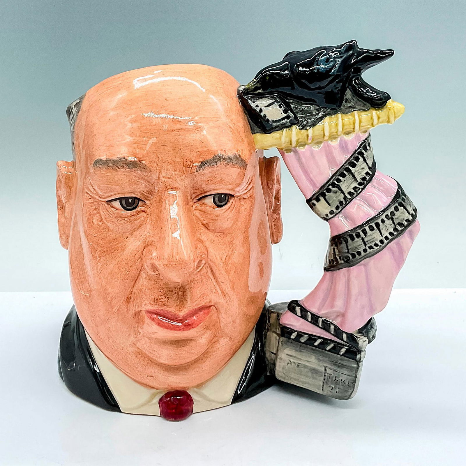Alfred Hitchcock D6987 (Colorway) - Large - Royal Doulton Character Jug: D6987; featuring a pink shower curtain handle. Royal Doulton backstamp. Artist: David B. Biggs Issued: c. 1995 Dimensions: 7.5"L x 6"W x 7"H Manufacturer: Royal Doulton Country of Origin: England