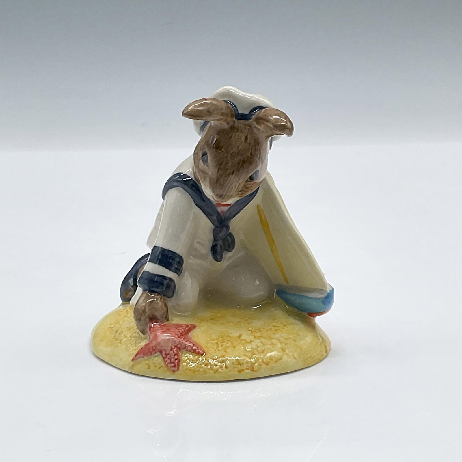 Sailor DB166 - Royal Doulton Bunnykins: Glossy porcelain. Features a bunny dressed in white and blue sailor uniform holding a boat by his side. Royal Doulton backstamp. Artist: Graham Tongue Issued: c. 1997 Dimensions: 2.75''L x 2.5''W x 2.