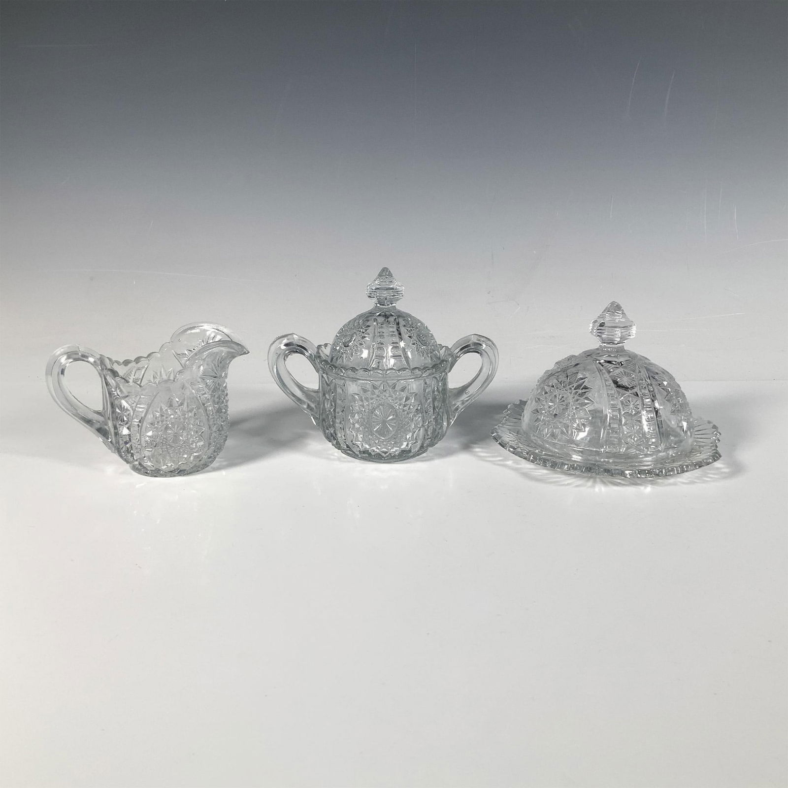 3pc Indiana Glass Oval Star Miniature Serveware: A stunning trio consisting of a butter plate with lid, a creamer, and a sugar bowl with lid, all featuring a star and geometric cut pattern. Butter plate with lid: 5.25"W x 3.5"H. Creamer: 4"L x 2.5"W