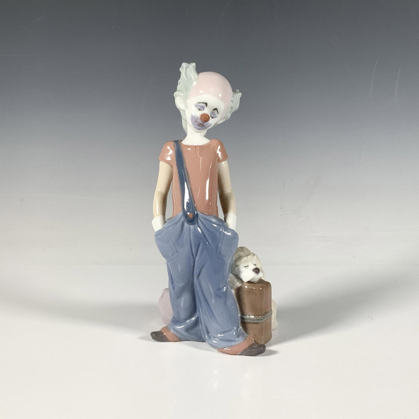 Destination Big Top 1006245 - Lladro Porcelain Figurine: A glossy figure of a sad looking clown with his dog and luggage beside him. 1996 Event Piece. Lladro backstamp. Artist: Juan Huerta Issued: 1996 Dimensions: 3.75"L x 3.5"W x 8"H Manufacturer: Lladro