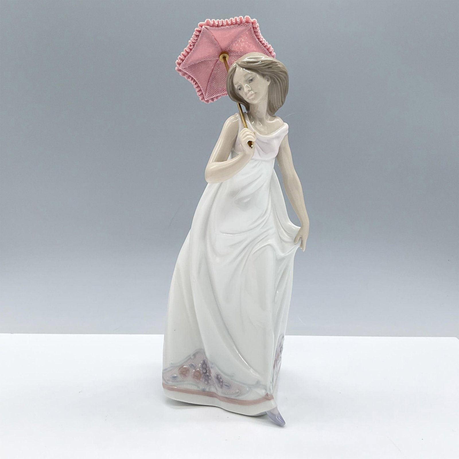 Afternoon Promenade 1007636 - Lladro Porcelain Figurine: Glossy porcelain, features a delicate lady dressed in a white dress holding a pink parasol. Lladro backstamp. Artist: Jose Puche Issued: 1995 Dimensions: 4.25"L x 4.25"W x 11"H Manufacturer: Lladro C