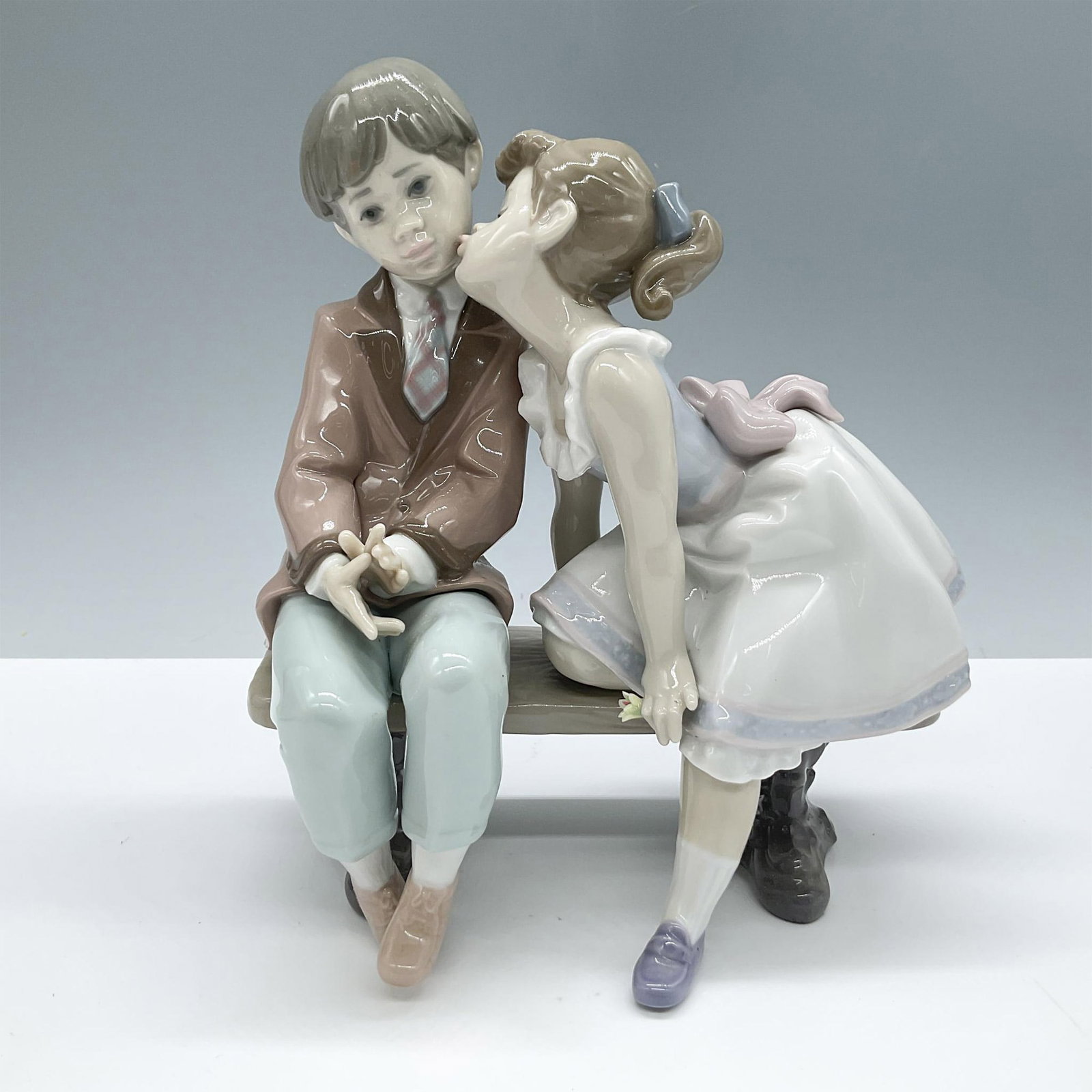 Ten & Growing 1007635 - Lladro Porcelain Figurine: Glazed porcelain figural of a girl about to kiss a boy on the cheek as they both sit on a bench. Lladro backstamp. Artist: Francisco Polope Issued: 1995 Dimensions: 7.25"L x 4.5"W x 7.75"H Manufacture