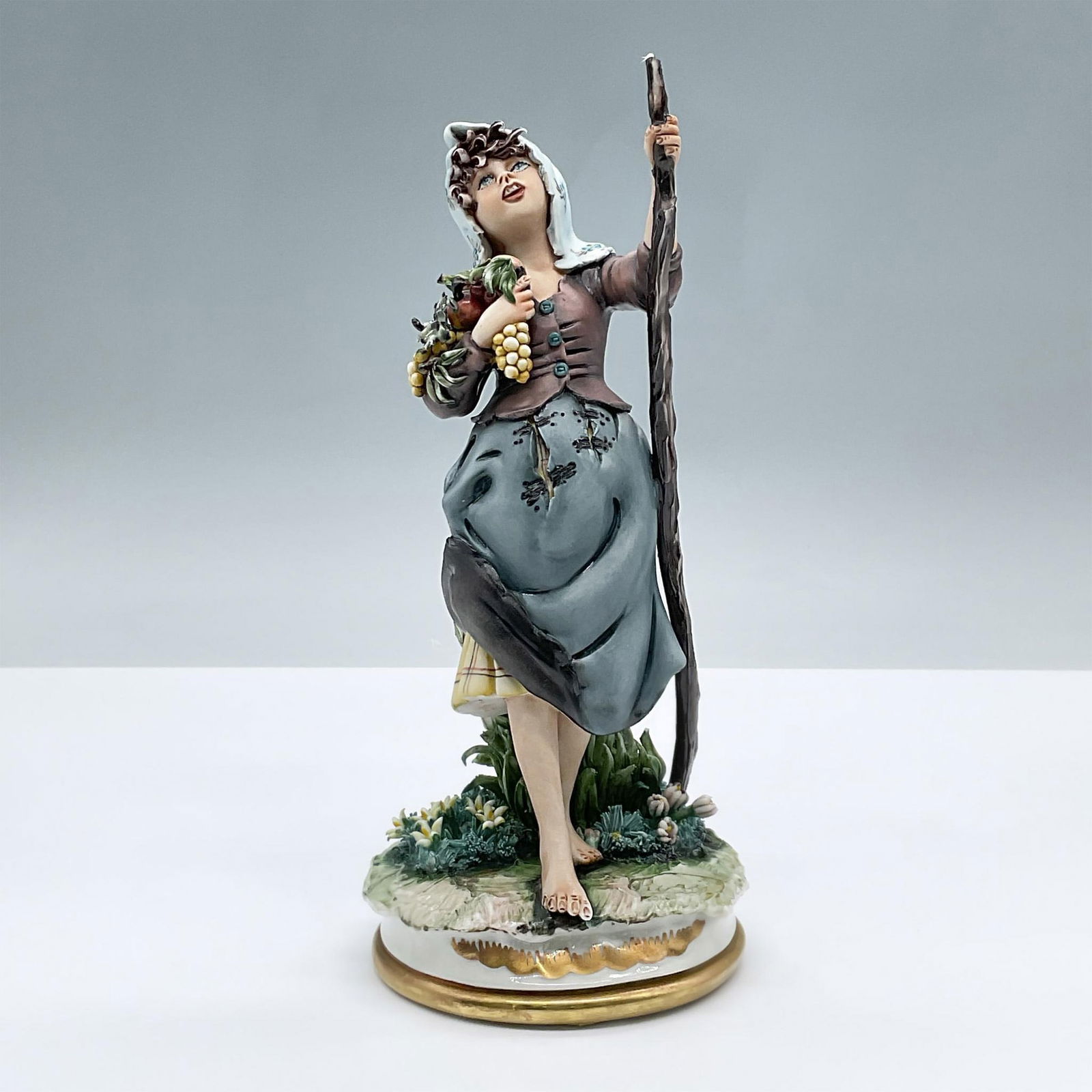 Capodimonte Porcelain Figurine, Peasant Woman with Fruit: Porcelain figure of a woman fashioned in peasant clothes with a walking stick, carrying a bundle of fruit, The base features gold gilded details. Signed by Artist. Capodimonte backstamp. Dimensions: 3