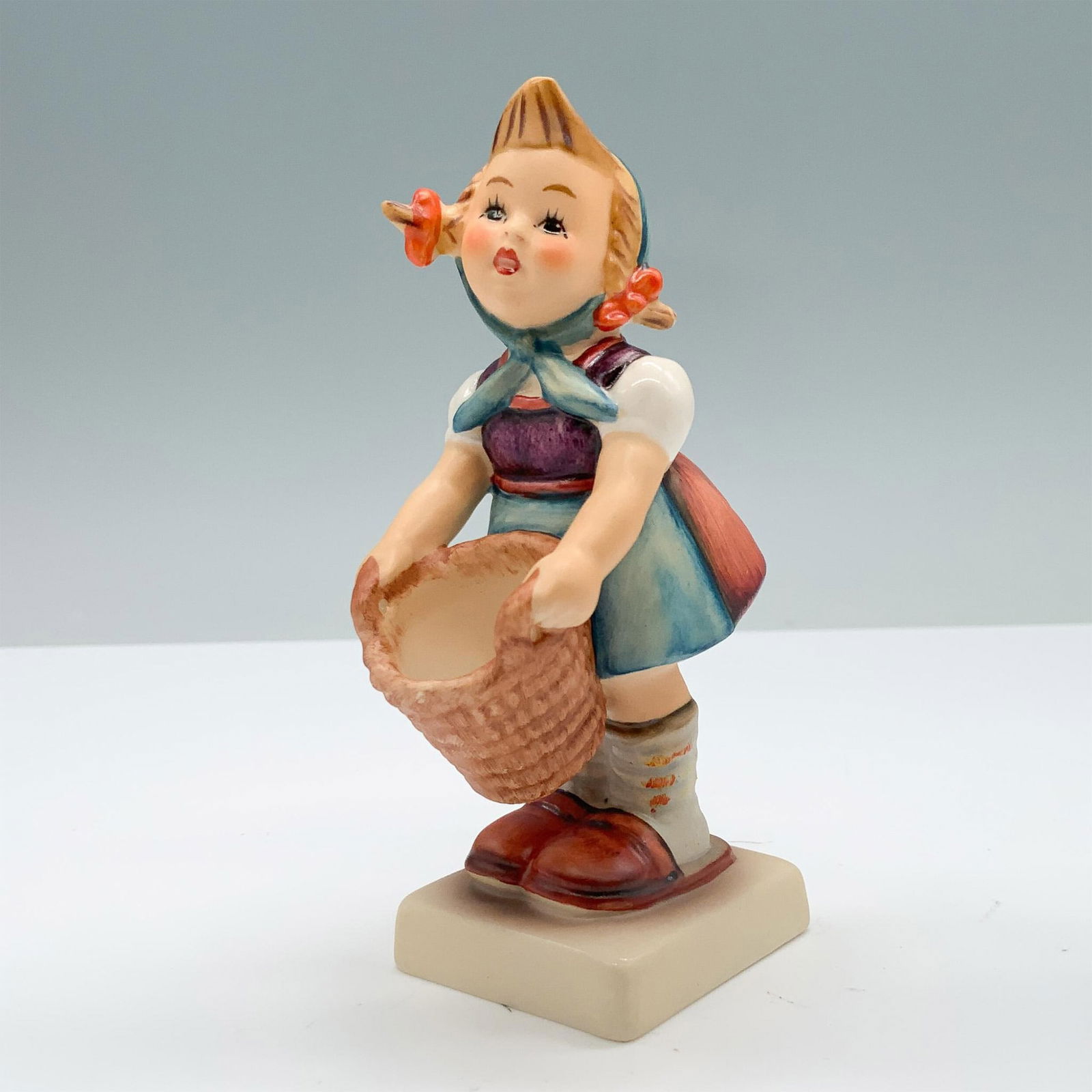 Goebel Hummel Figurine, Little Helper: Hand painted porcelain showcasing a young girl in a red dress with a blue bonnet, holding a basket in her hands. Goebel backstamp. Hummel-Hummel impressed. HUM73. This item has its original box. Box m