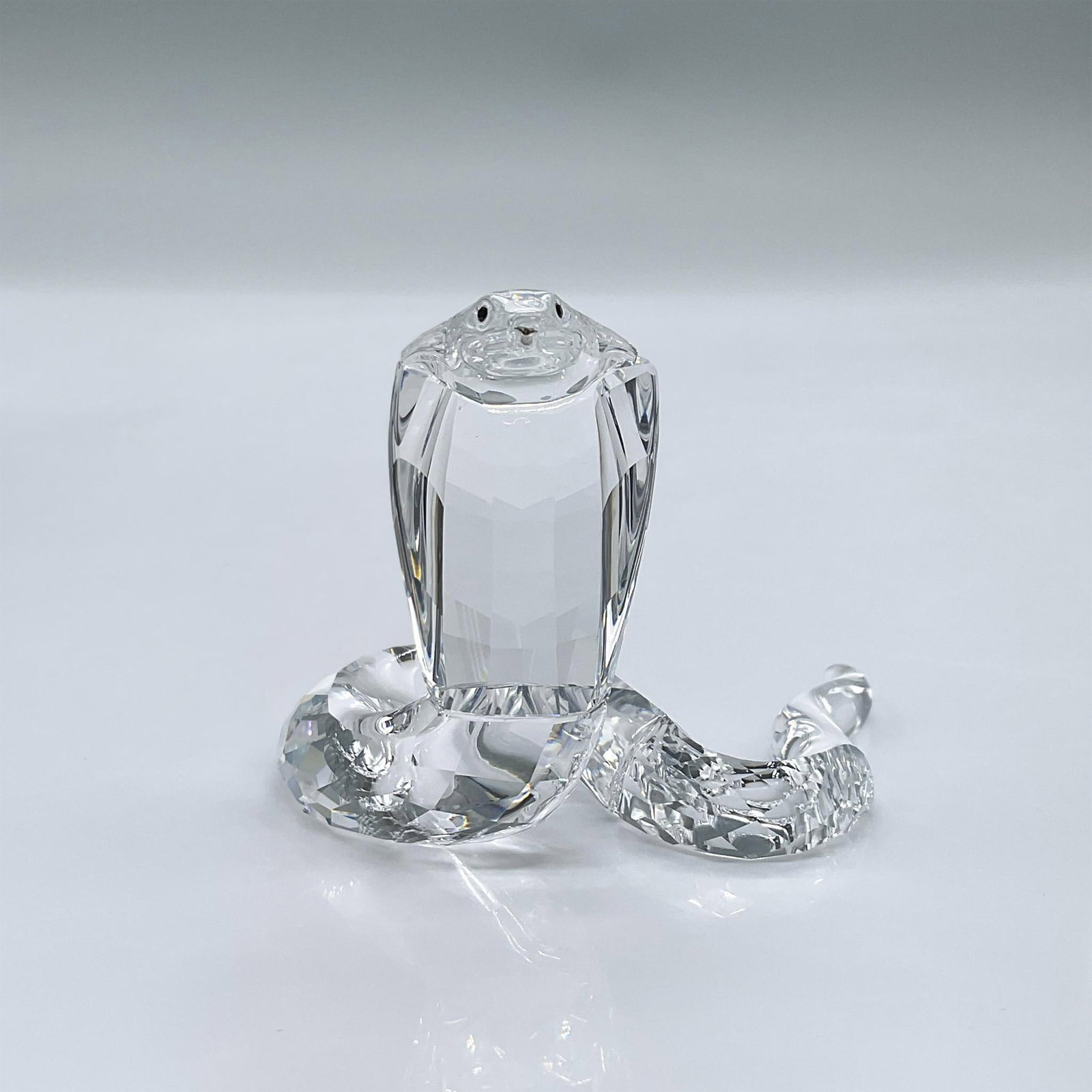 Swarovski Crystal Figurine, Cobra Snake: Made of clear facetted crystal with black eyes and a rhodium forked tongue. This item has its original box measuring 5"L x 5"W x 4"H. Swarovski acid mark. 243979. Artist: Heinz Tabertshofer Issued: 20