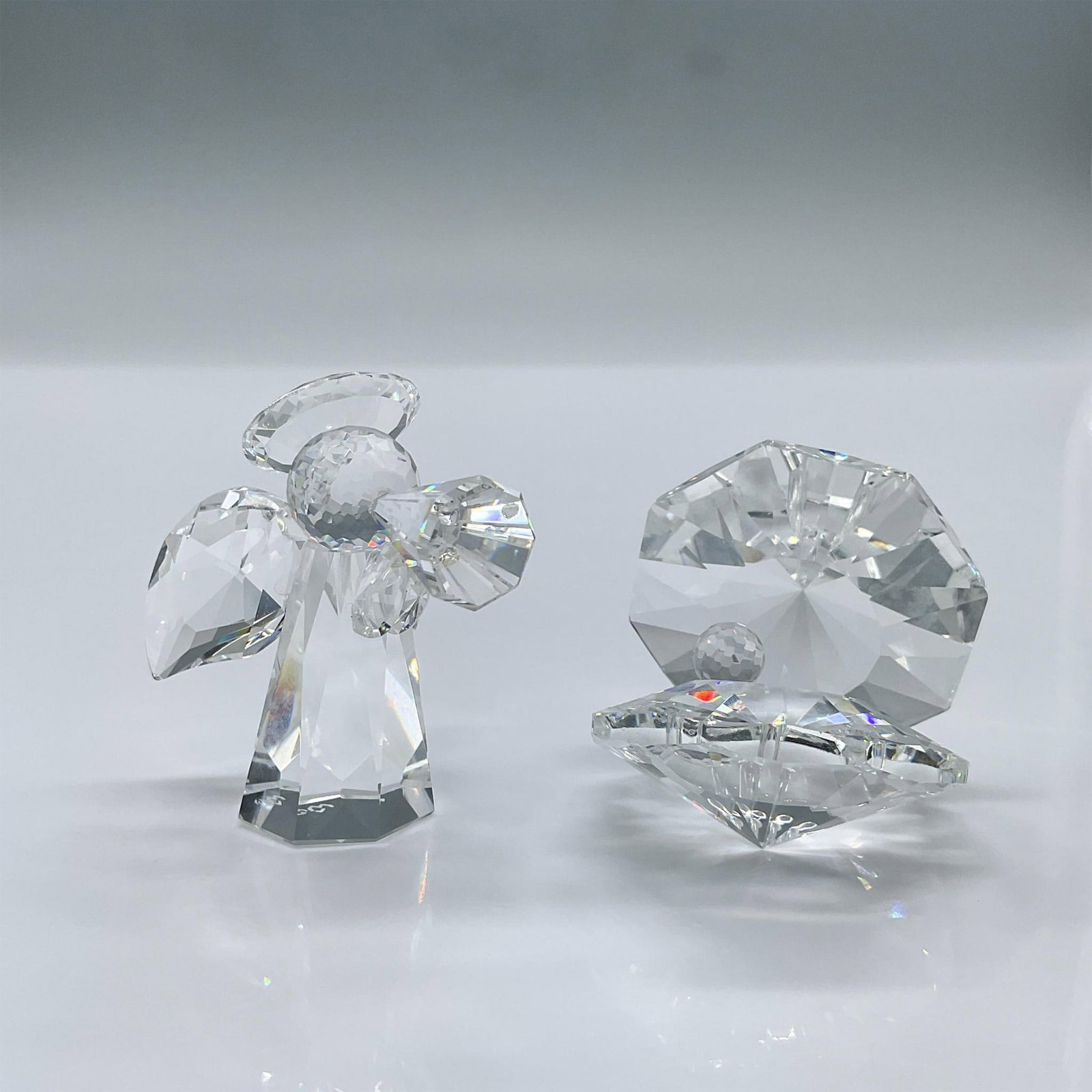 2pc Crystal Angel and Clam Figurines: This is a set of 2 elegant clear crystal figurines of an angel with a trumpet, and a clam with a pearl. Largest measures 2"L x 1.5"W x 2.25"H. The angel has a makers mark acid stamp. Dimensions: See D