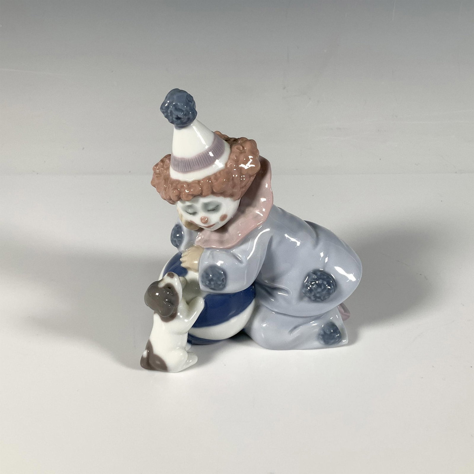 Pierrot with Puppy & Ball 1005278 - Lladro Porcelain Figurine: A glossy figure of a young clown leaning over a ball and staring at a dog. Lladro backstamp. Artist: Jose Puche Issued: 1985-2007 Dimensions: 4.25"L x 2.75"W x 4.75"H Manufacturer: Lladro Country of