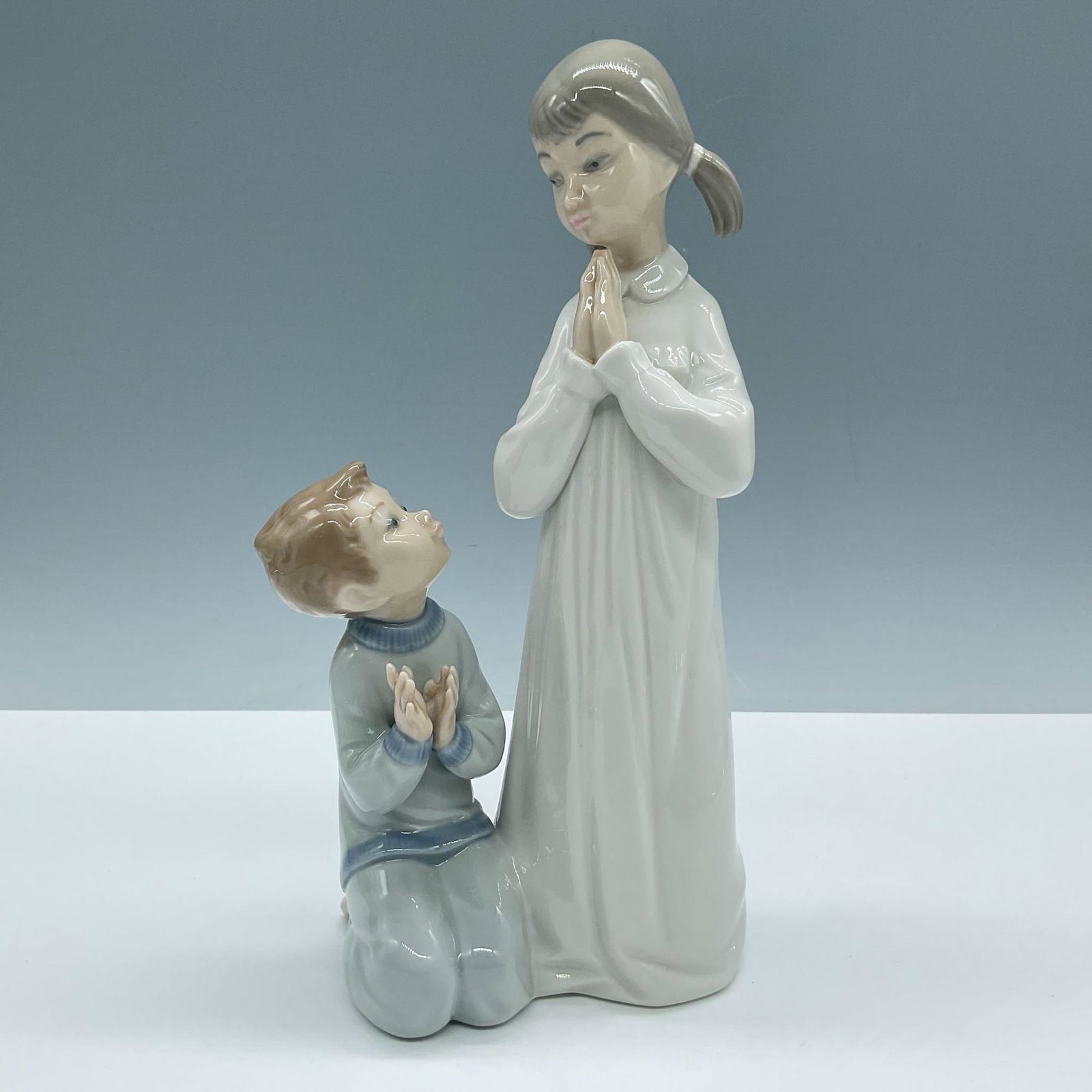 Lladro Porcelain Figurine, Teaching to Pray 10004779: A glossy figure of a sister teaching her brother how to pray. Lladro backstamp. Artist: Vincente Martinez Issued: 1971-1997 Dimensions: 4.5''L x 4''W x 8.75''H Manufacturer: Lladro Country of Origin: