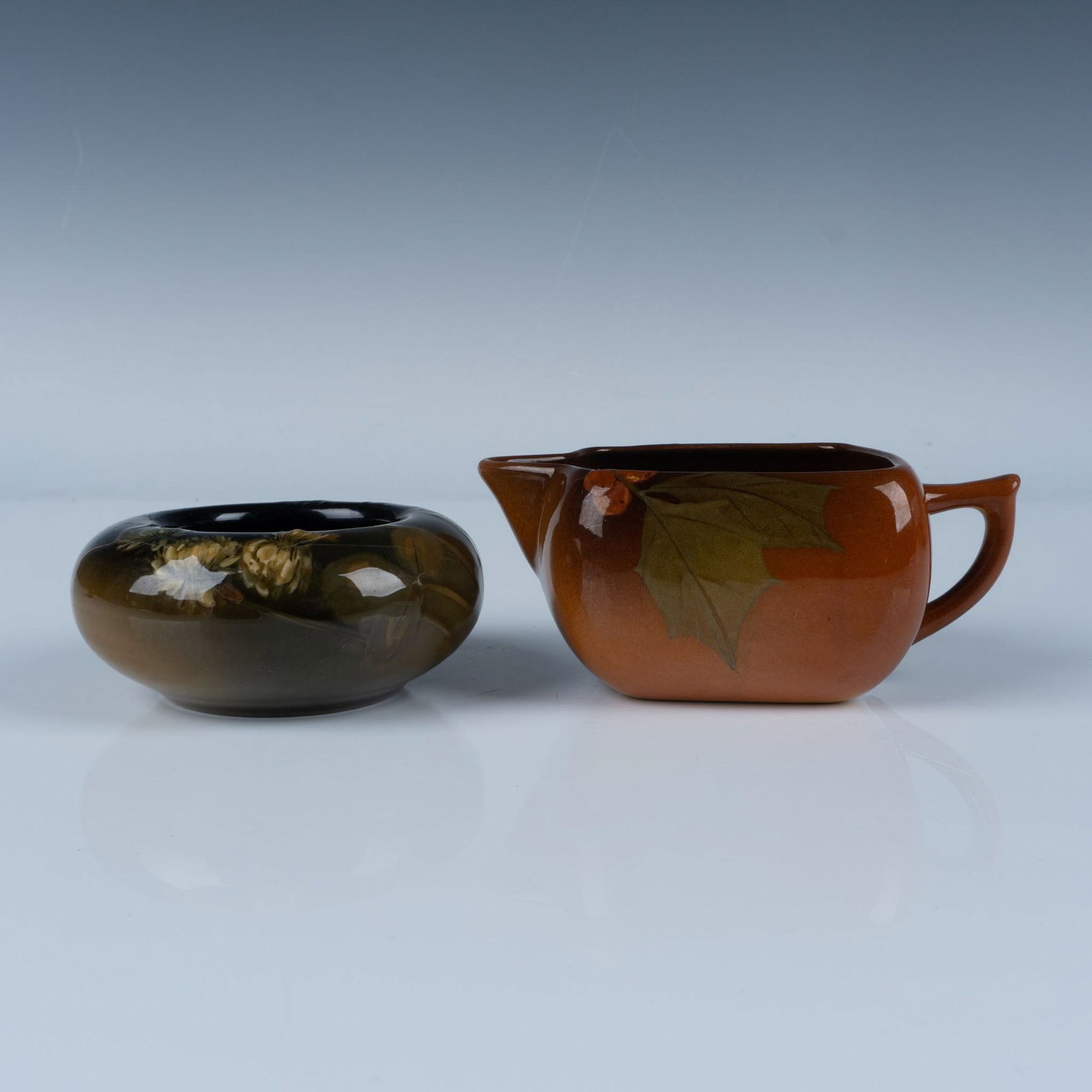 2pc Rookwood Pottery Botanical Bowl and Creamer: High glazed, collection includes a small bowl with hand painted white clovers, and a creamer with red holly berries. Rookwood Pottery impressed stamp, and marked artists. Bowl: Marked VI; Shape 1142; 