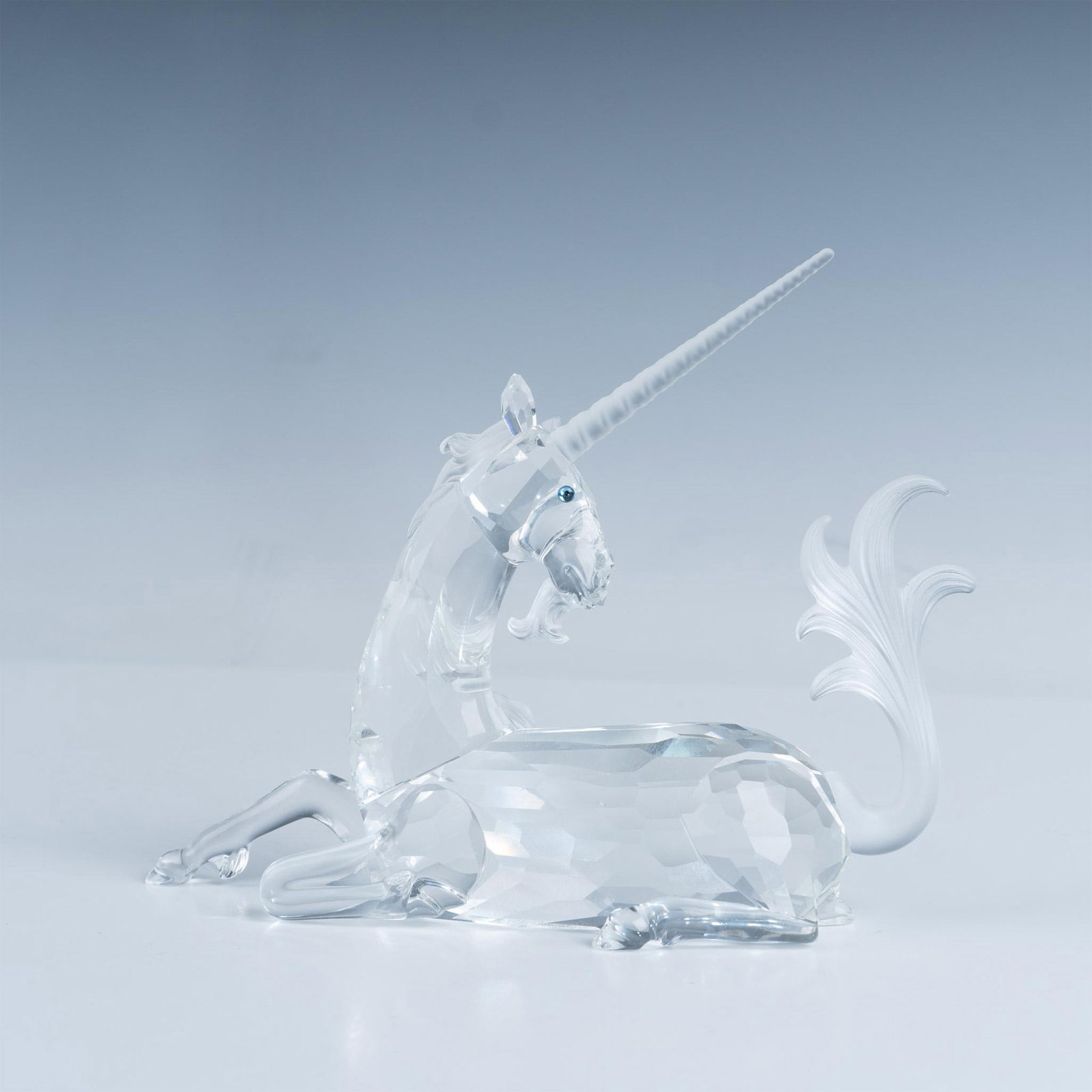Swarovski Crystal Figurine, Unicorn 191727: A graceful and elegant unicorn sits on all fours, its head turned in thoughtful observation, showcasing mesmerizing blue crystal eyes and a frosted tail. Swarovski backstamp. This item has its origina