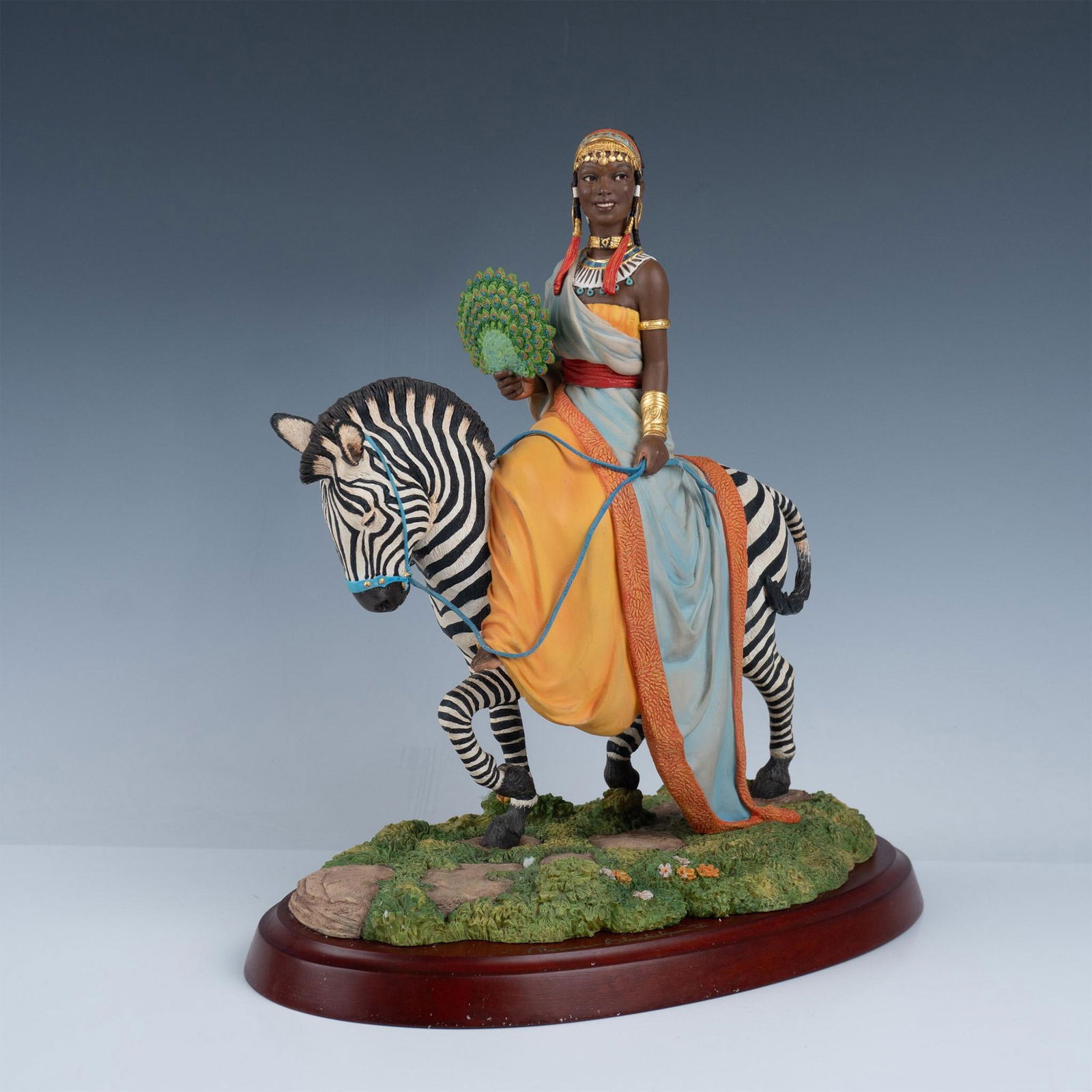 Thomas Blackshears Ebony Visions, The African Queen: This is a wonderfully elaborate sculpture of a beautiful African woman riding on a zebra. She is adorned in gold, and fashioned in an orange dress and light blue drapey robe with orange textured detai