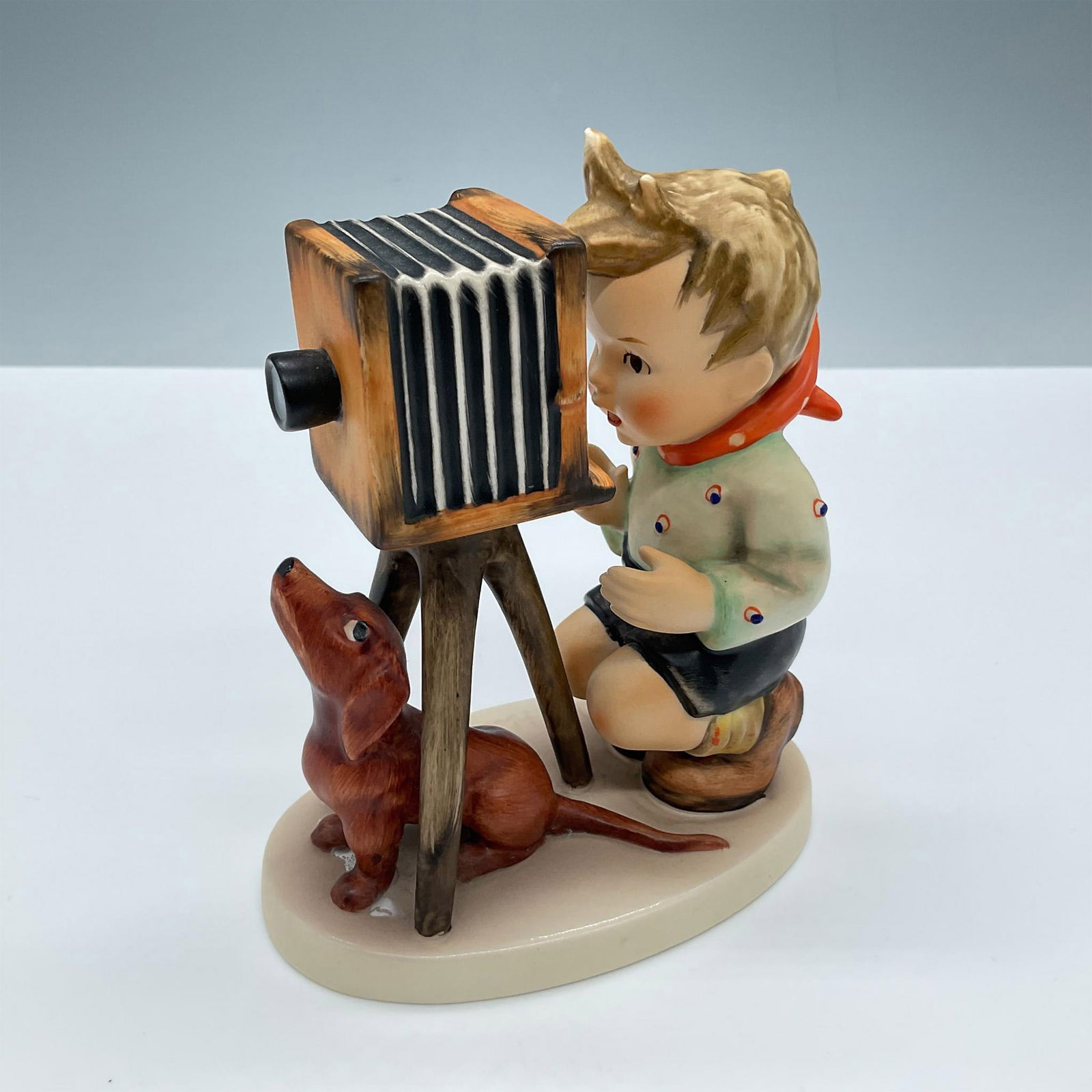 Goebel Hummel Figurine, The Photographer: Hand painted in matte finish. A young boy takes photos with his pup by his side. Goebel Hummel backstamp. Issued: 20th century Dimensions: 4''L x 2.5''W x 5''H Manufacturer: Goebel Hummel Country of O